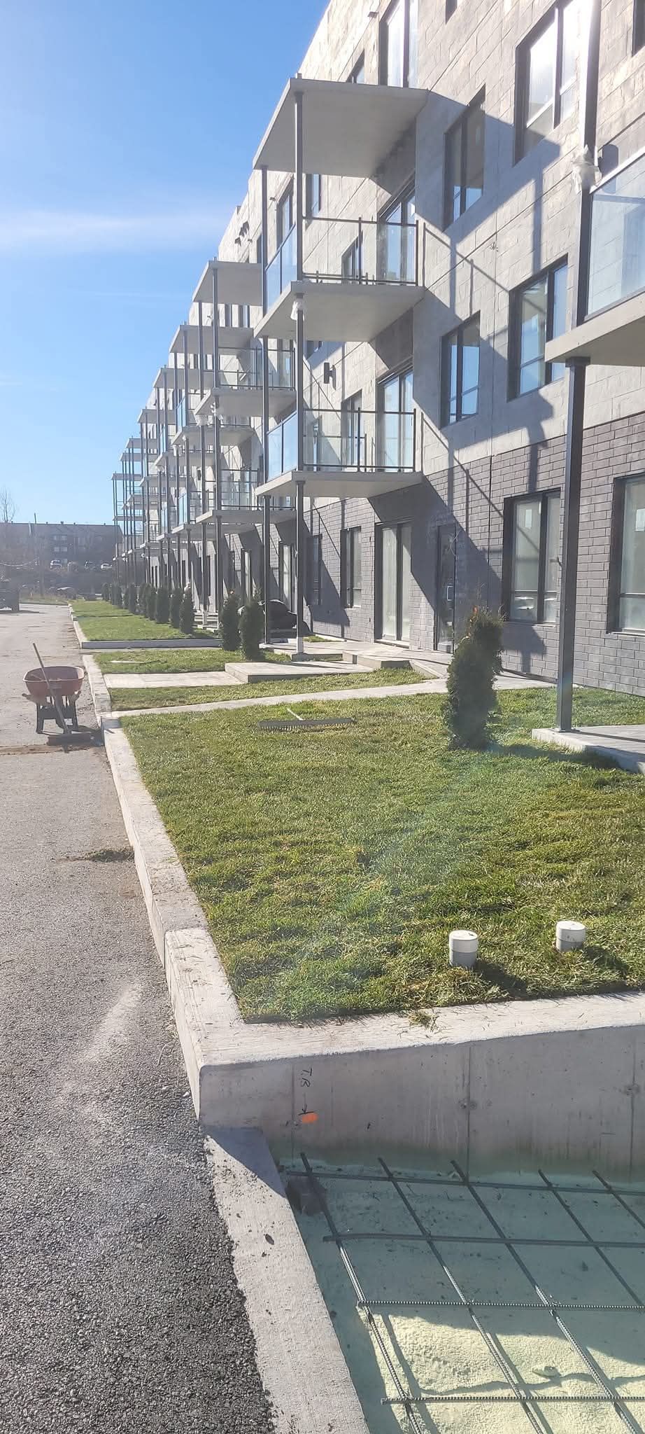 Vue extérieure d'un immeuble d'appartements moderne avec balcons, pelouse et chemin de gravier par une journée ensoleillée.
