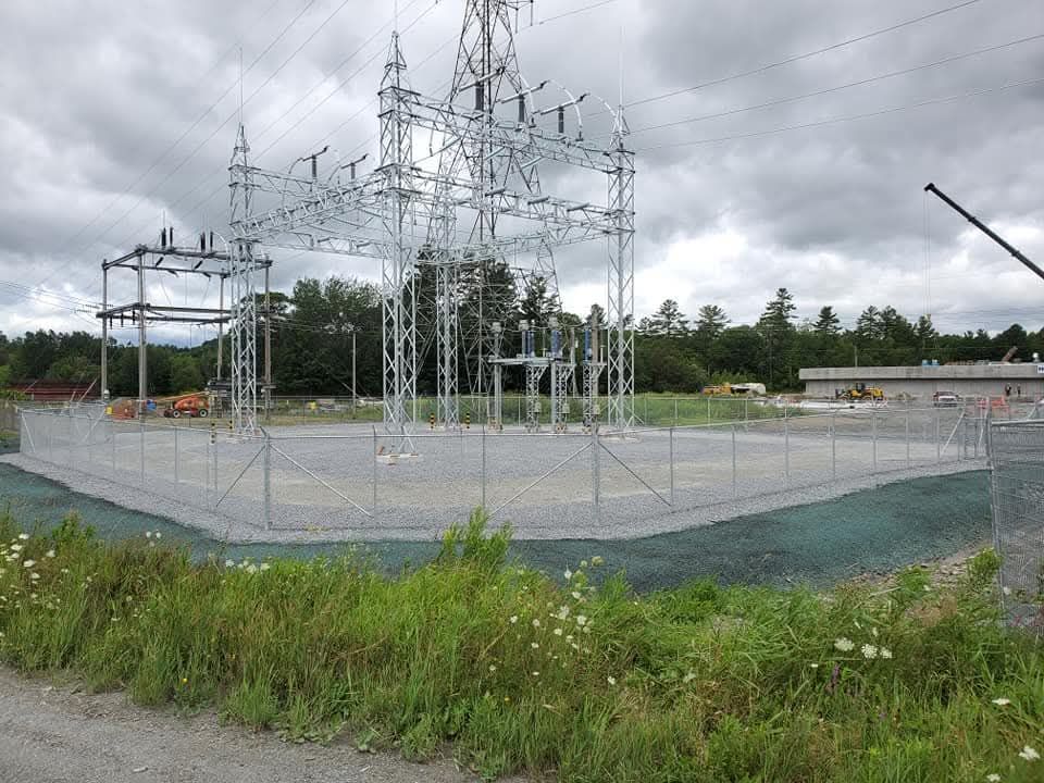 Poste de transformation électrique entouré d'une clôture en grillage, sous un ciel nuageux.