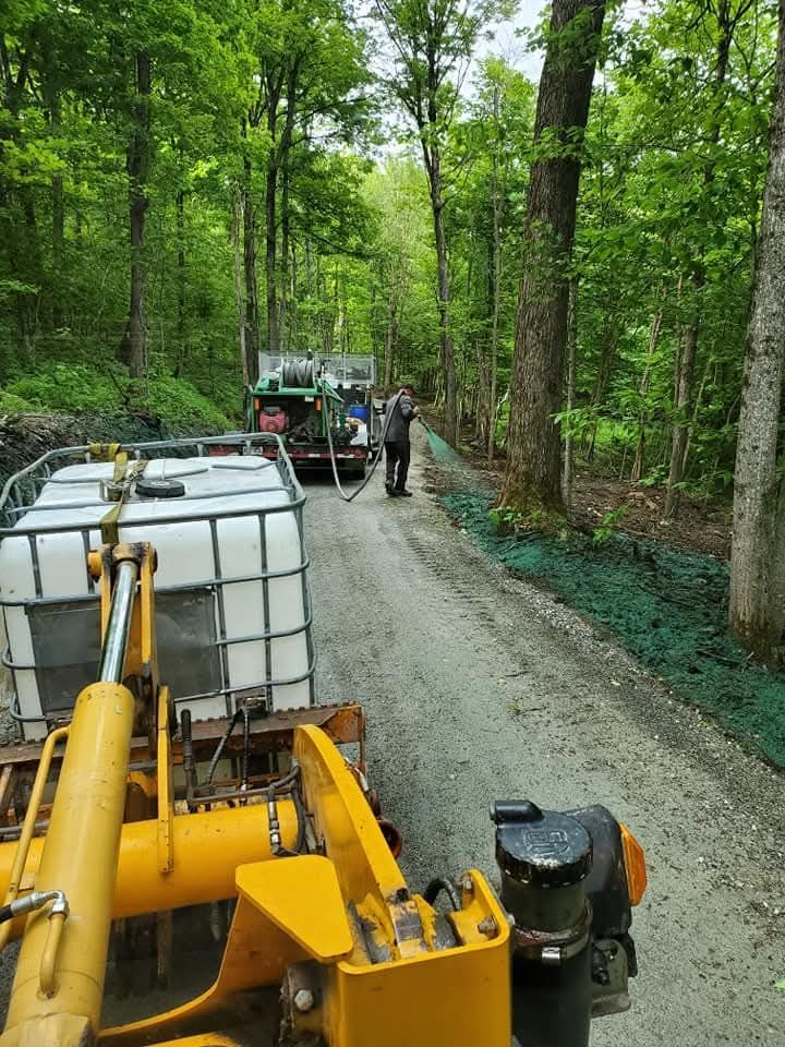 Un tracteur jaune remorque des citernes d'eau sur un chemin de gravier traversant une zone boisée ; une personne se tient à proximité.