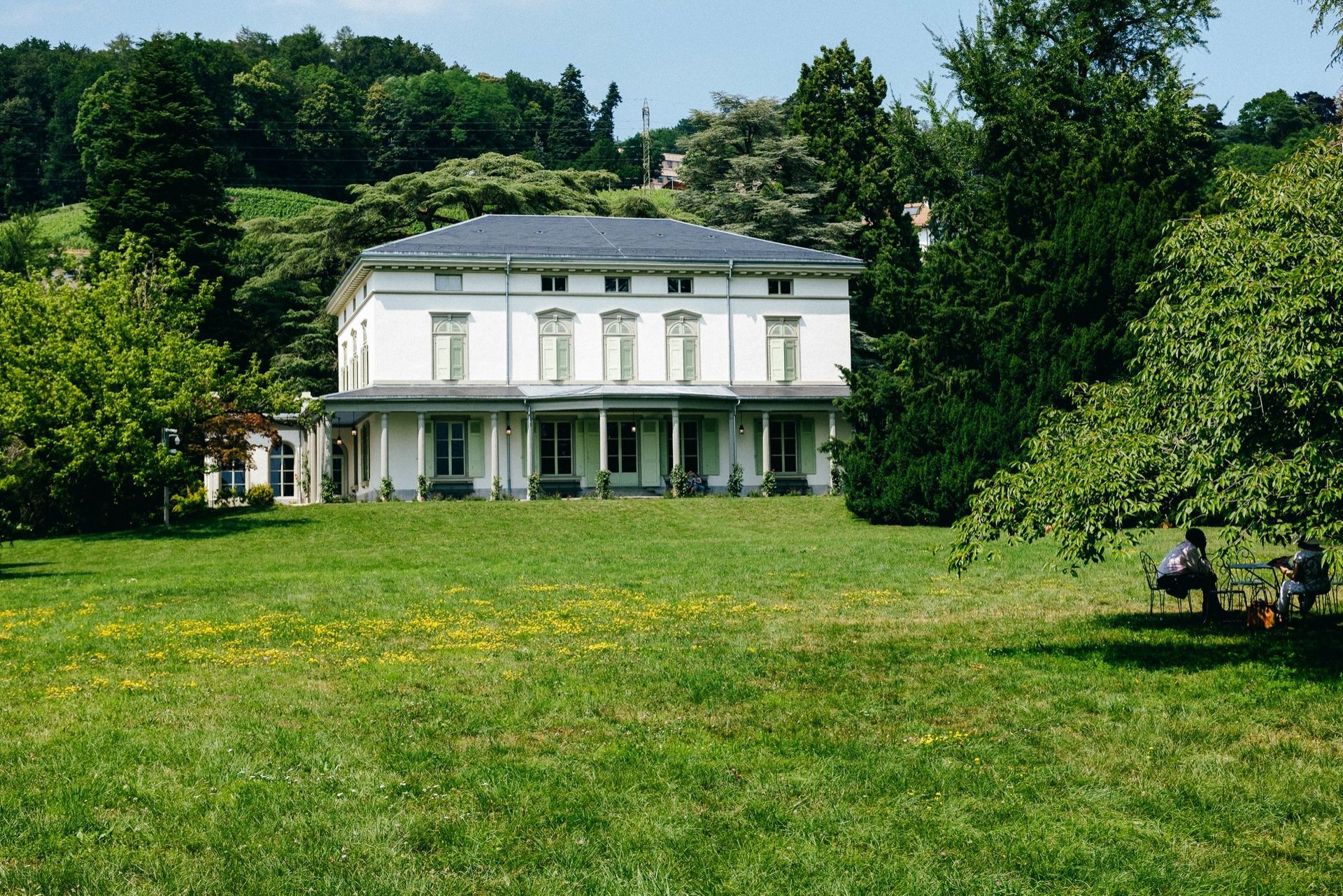 Un bâtiment blanc au toit sombre, entouré d'arbres et d'herbe verdoyants, par une journée ensoleillée. Deux personnes sont assises sur un banc.