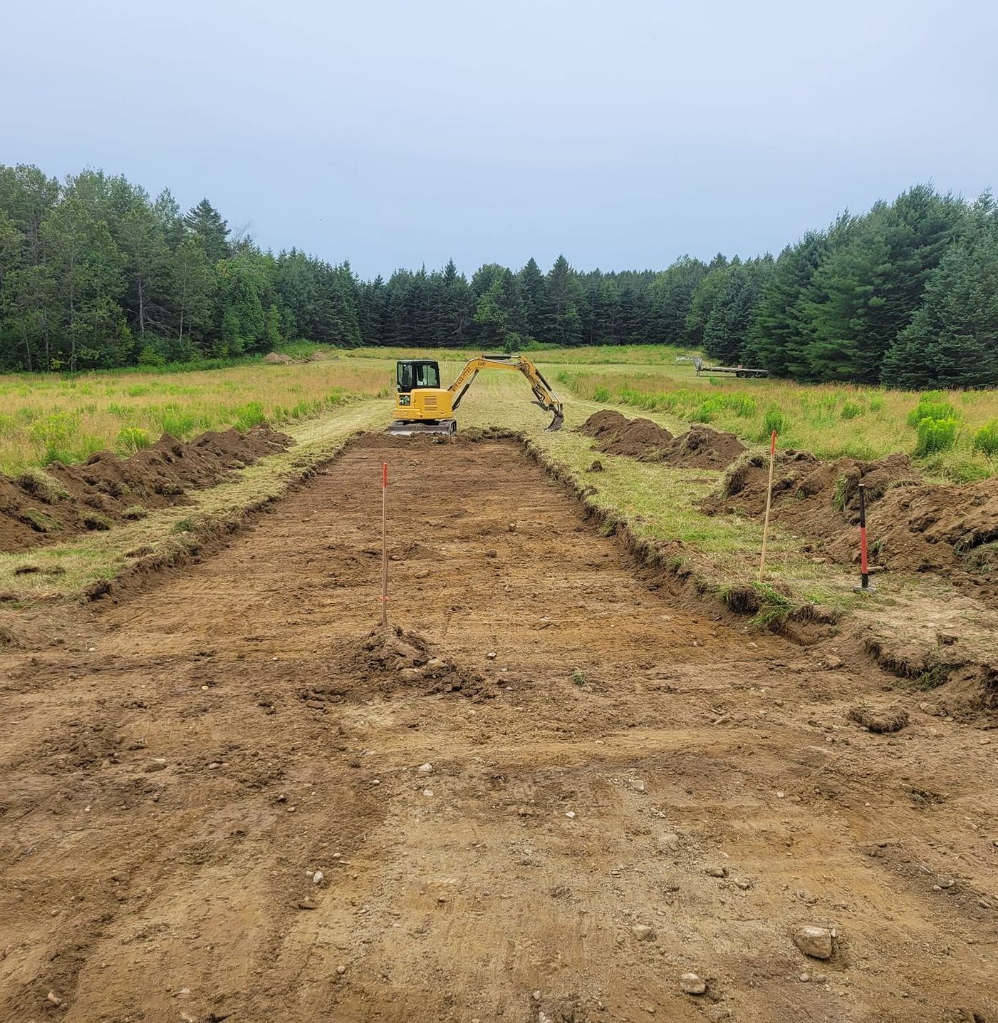 Chantier de fouilles avec une petite excavatrice, creusant des tranchées dans un champ, arbres en arrière-plan, ciel couvert.
