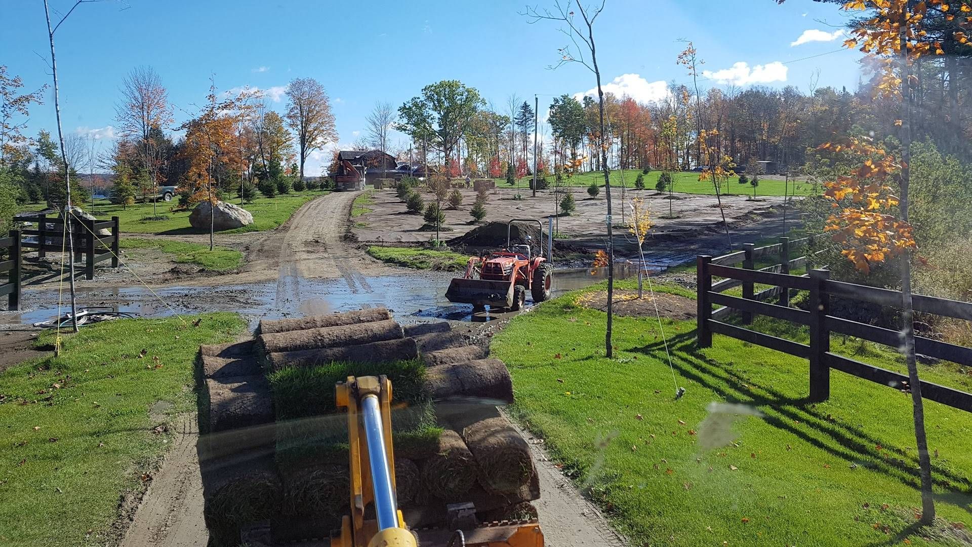Un tracteur déplace des rouleaux de gazon dans le cadre d'un projet d'aménagement paysager, avec un chemin boueux, des arbres et un ciel bleu en arrière-plan.