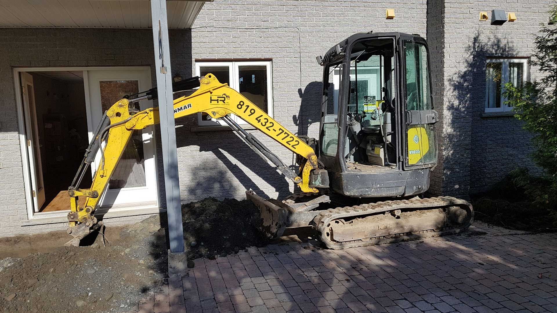 Une pelleteuse jaune enlève de la terre à côté d'un bâtiment à façade en briques et porte blanche.
