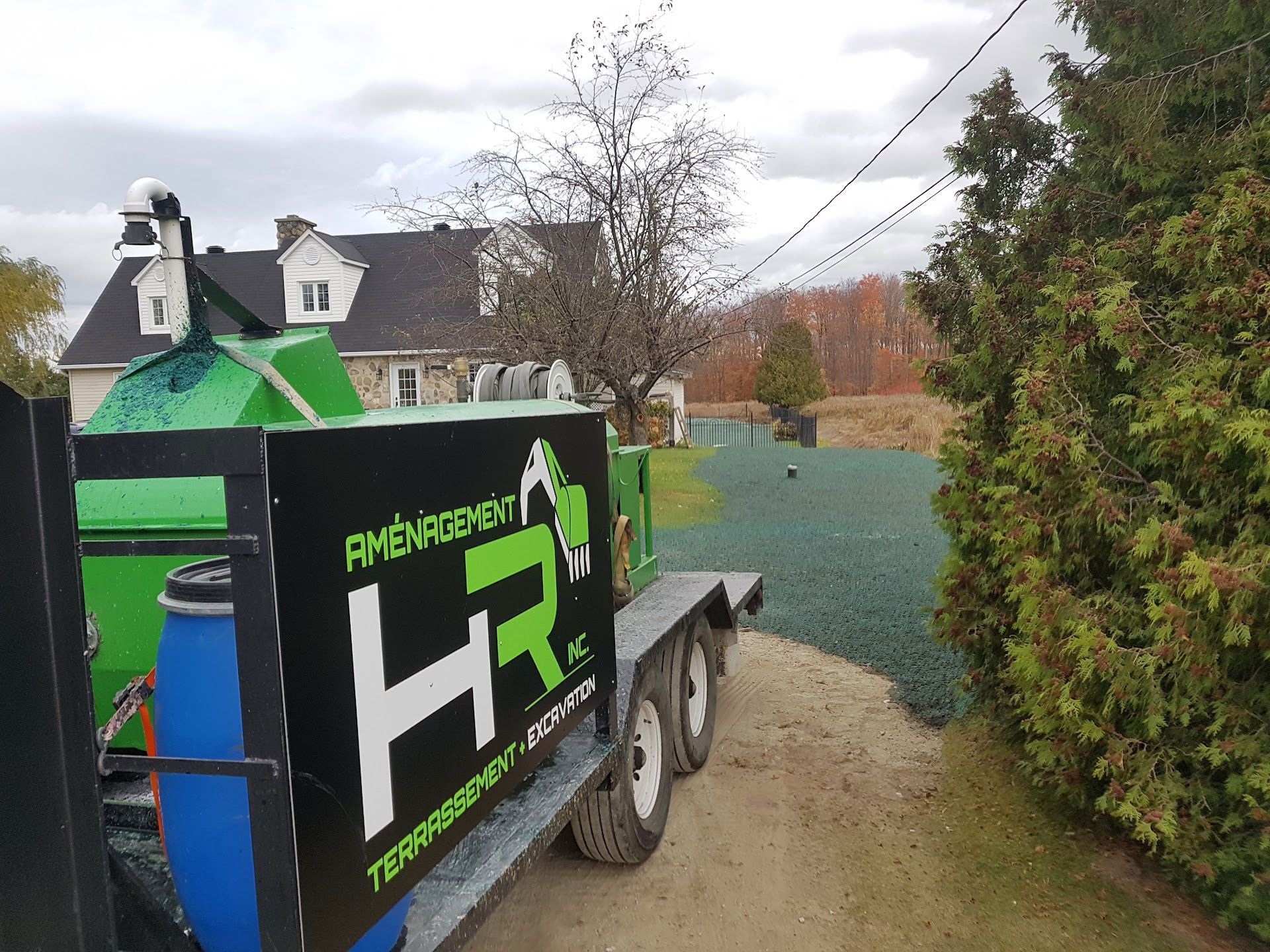 Broyeur de branches vert avec le logo « HR Amenagement » sur une remorque, près d'une maison et d'arbres.