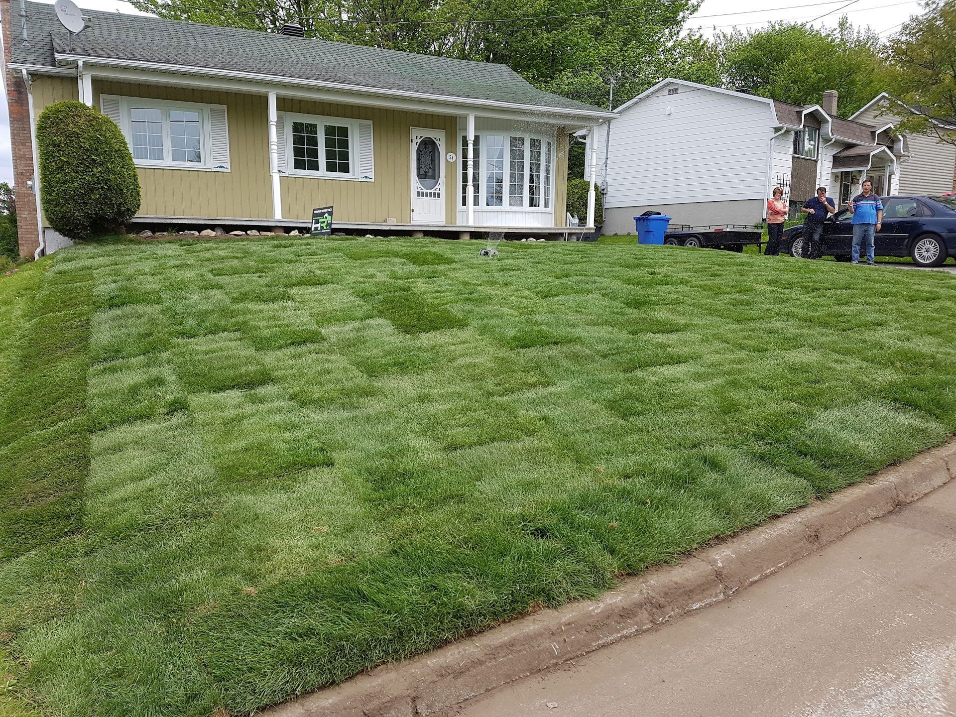 Maison avec pelouse fraîchement posée devant la maison. Des personnes se tiennent près d'une voiture. Pelouse verte, maison beige, poubelle bleue.