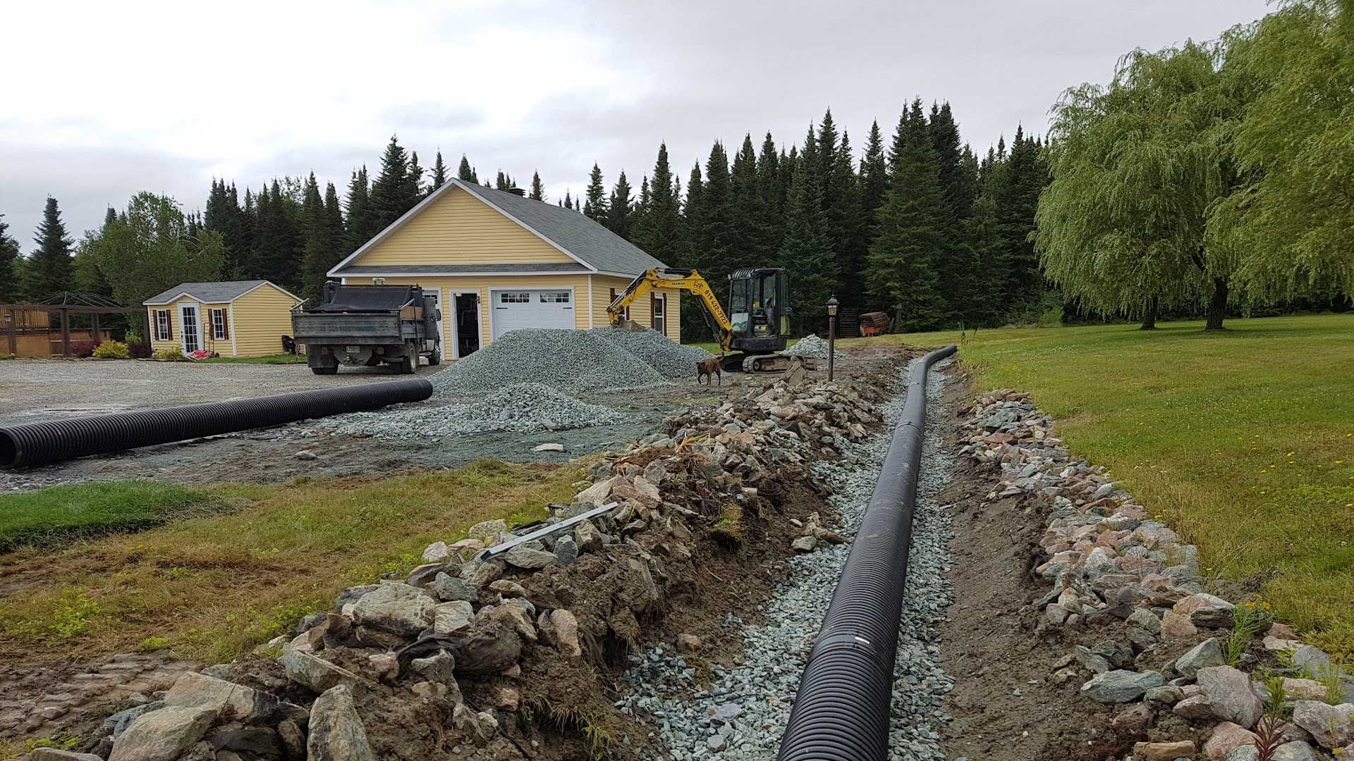 Tranchée creusée pour un tuyau de drainage dans une cour près d'un garage jaune et d'un petit bâtiment.