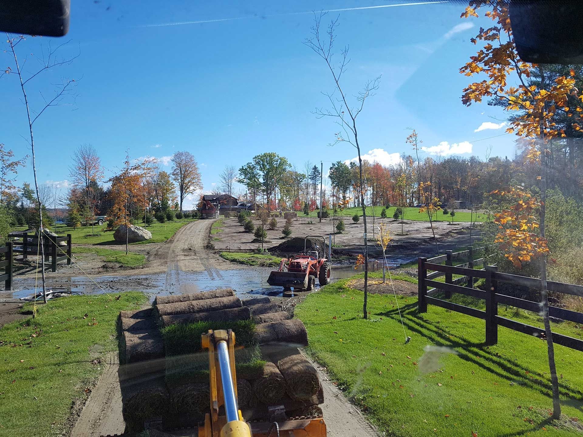 Un tracteur transporte des rouleaux de gazon sur un chantier de construction, avec des arbres et un ciel nuageux en arrière-plan.