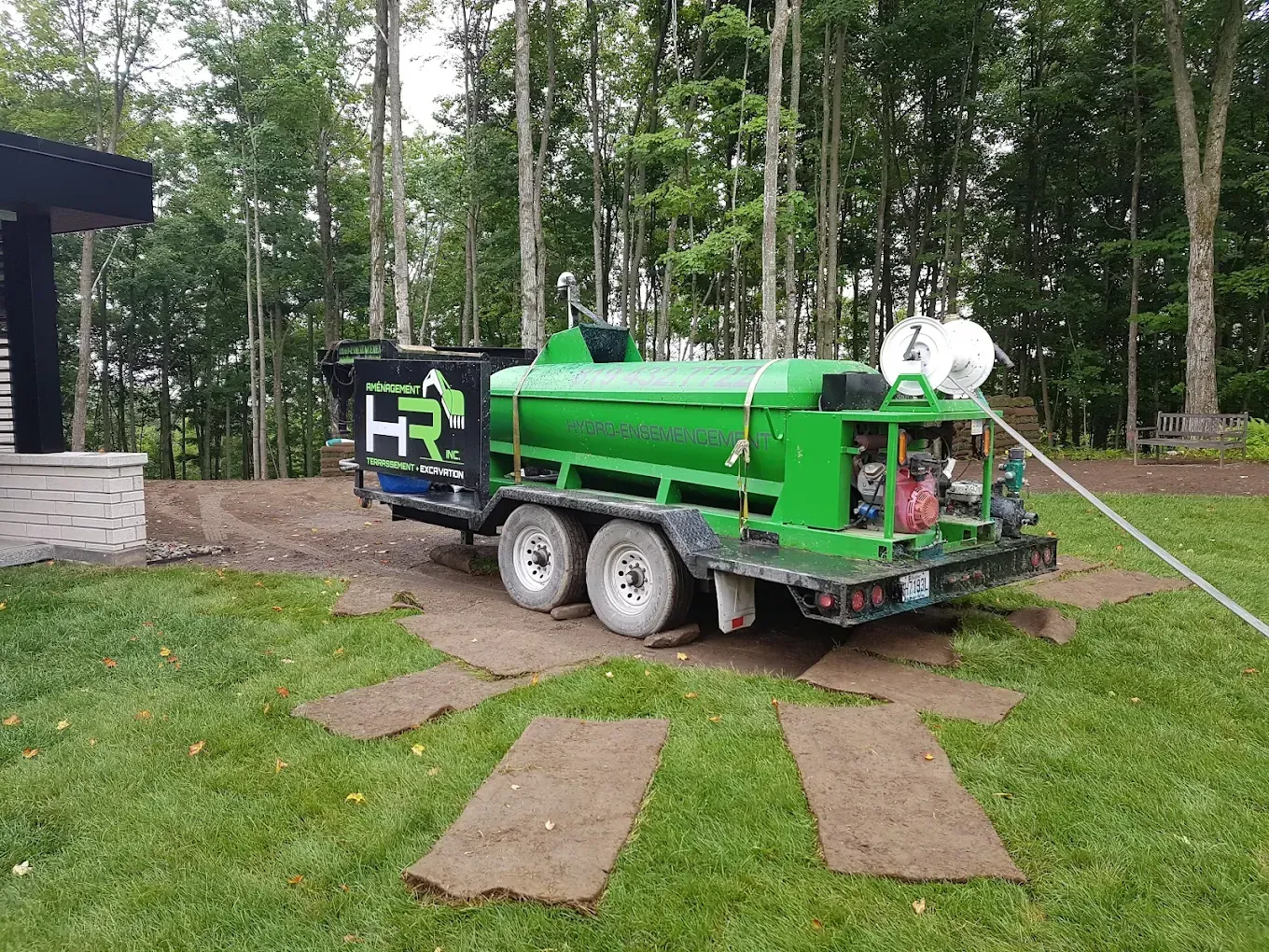 Caravane verte et noire sur pelouse avec des plaques de gazon. Arbres en arrière-plan, maison à gauche.
