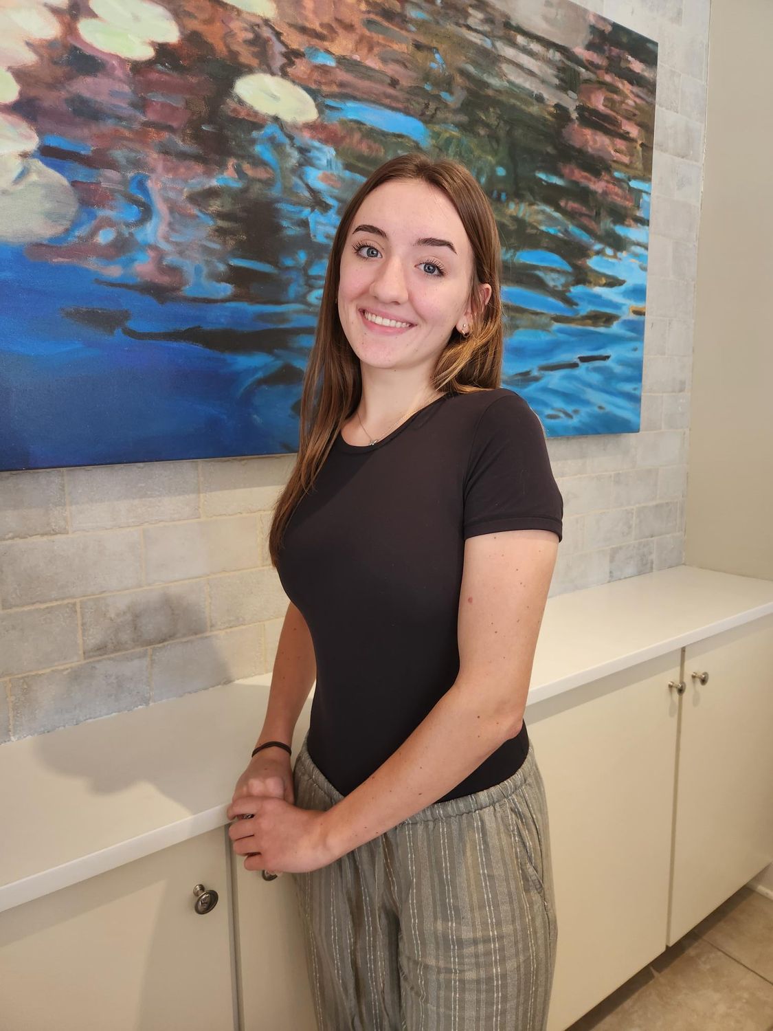 Woman in black shirt and striped pants smiles, standing by white cabinet under abstract blue art.