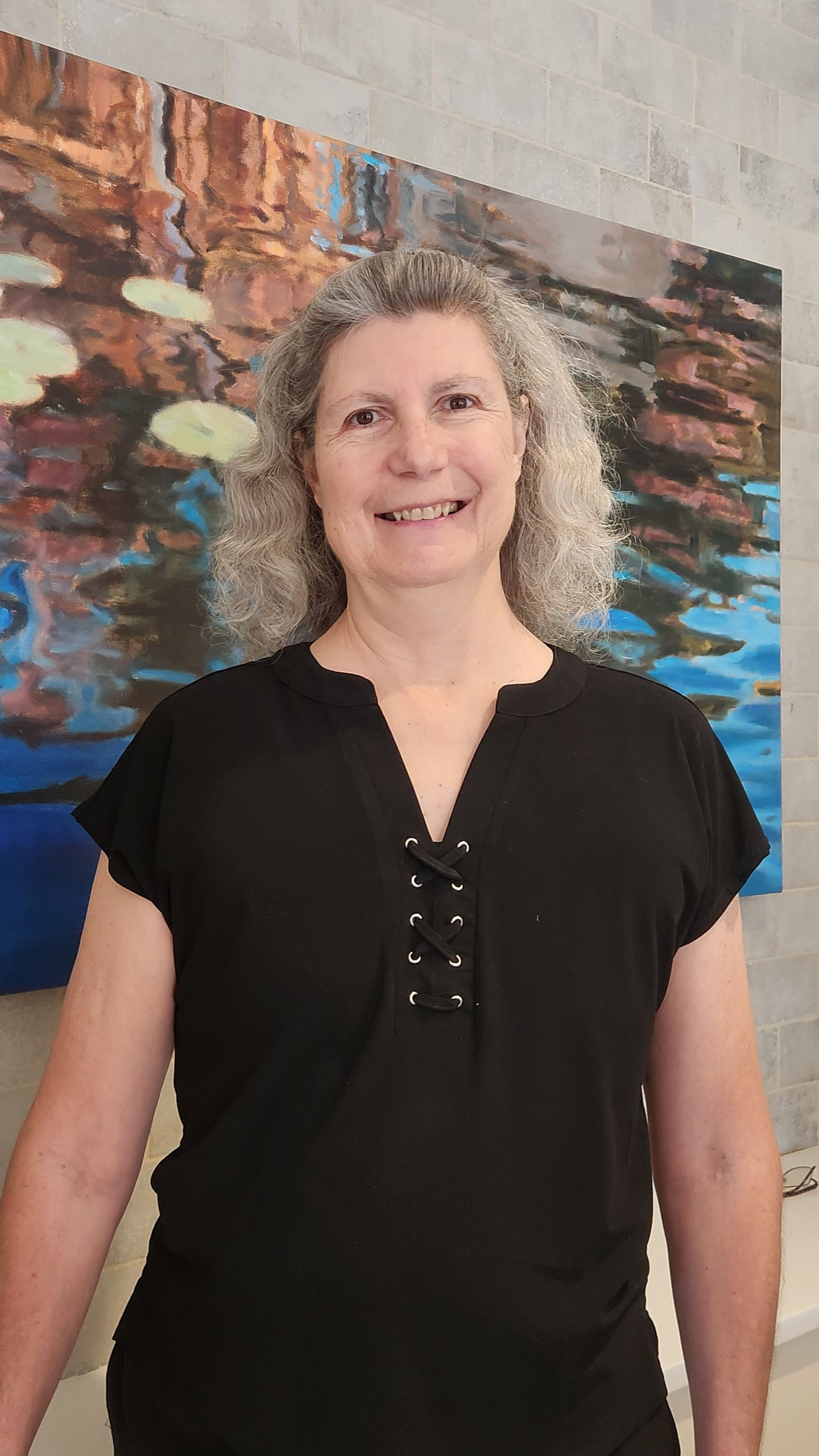 Woman with curly gray hair, black top, smiling in front of a painting with water reflection.