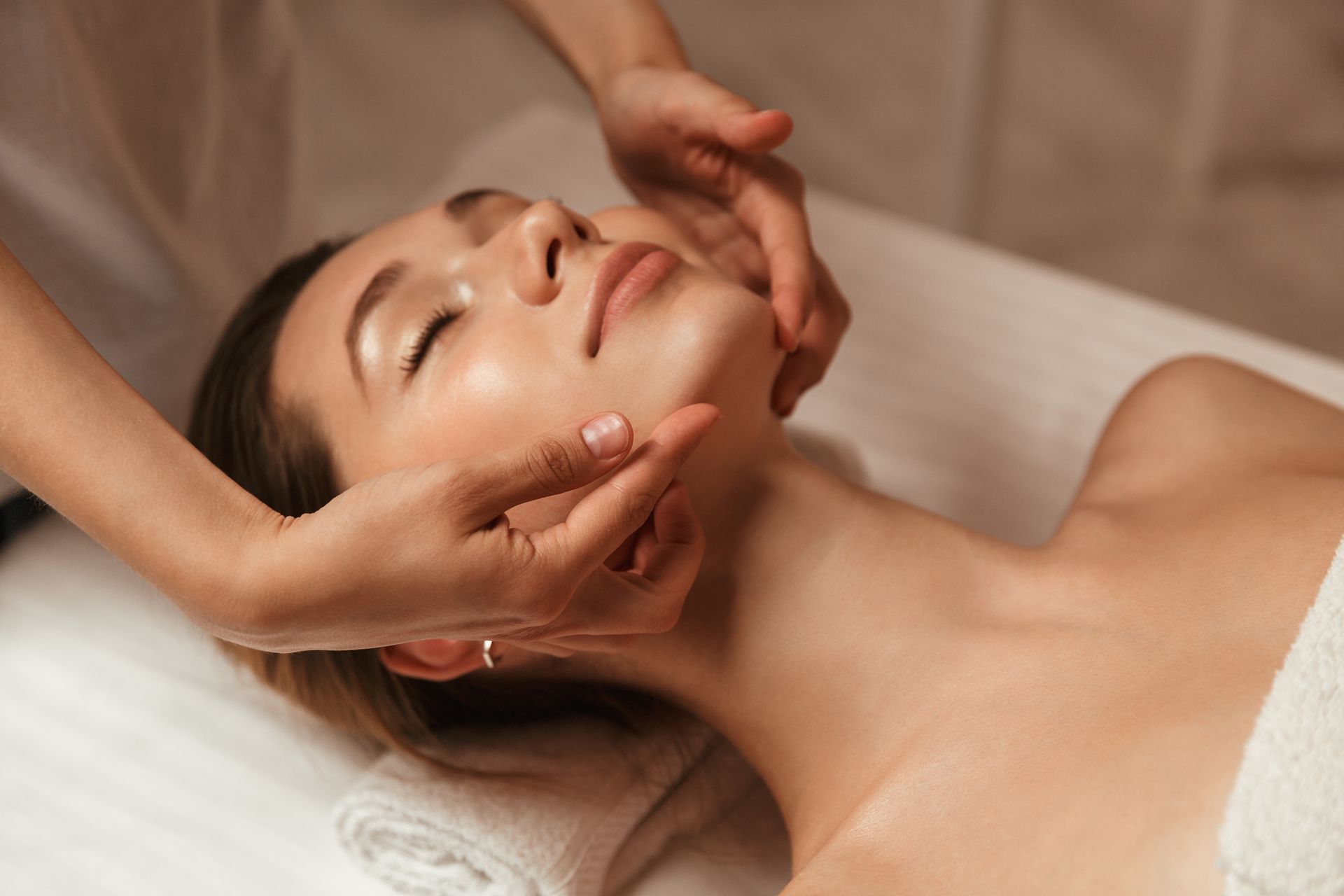 Woman receiving facial massage at a spa. Hands on her face, towel on chest, lying on a massage table.