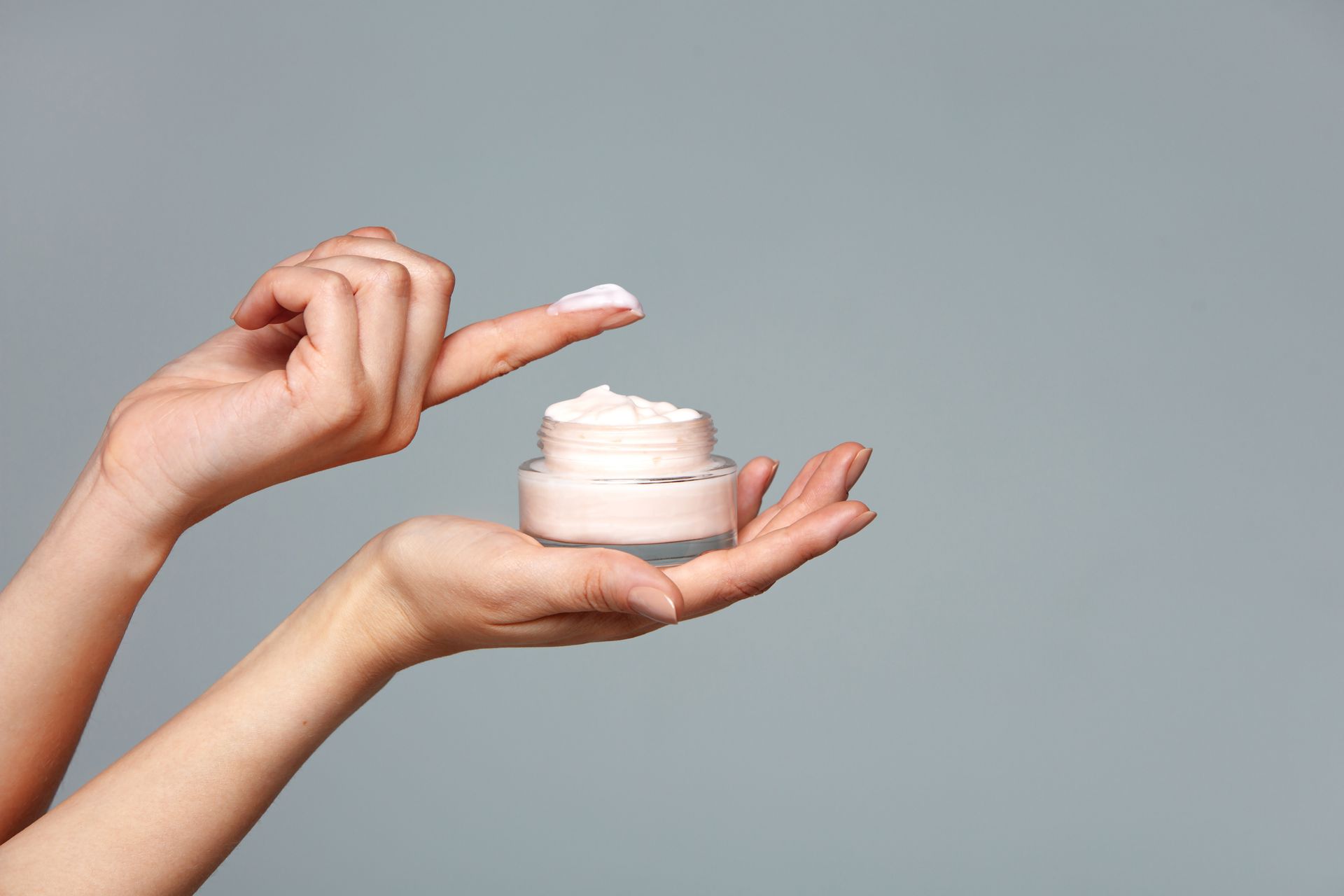 Hands holding a jar of cream, one finger has cream on it against a gray background.