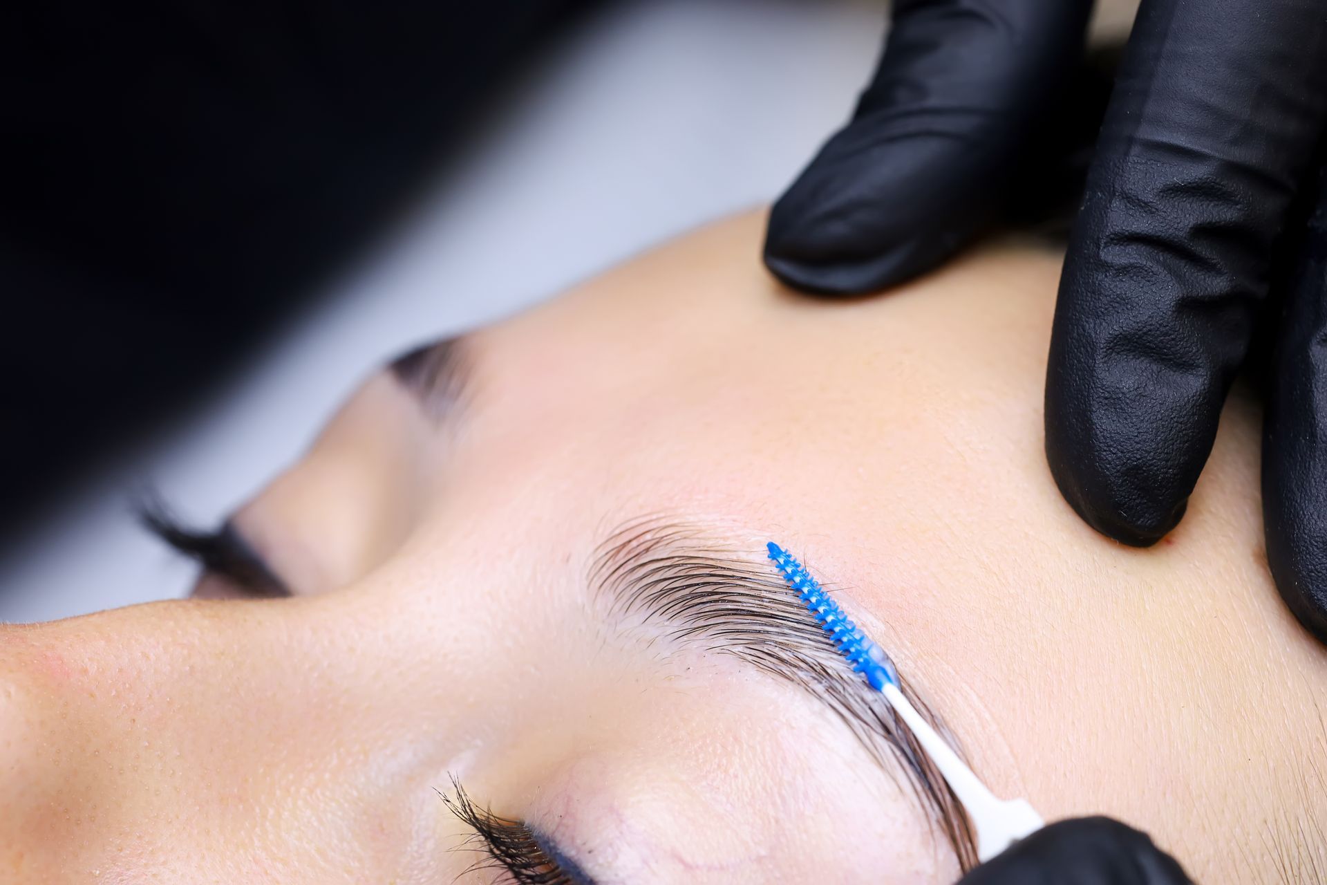 Close-up of eyebrow being groomed with a blue-tipped brush by a gloved hand.