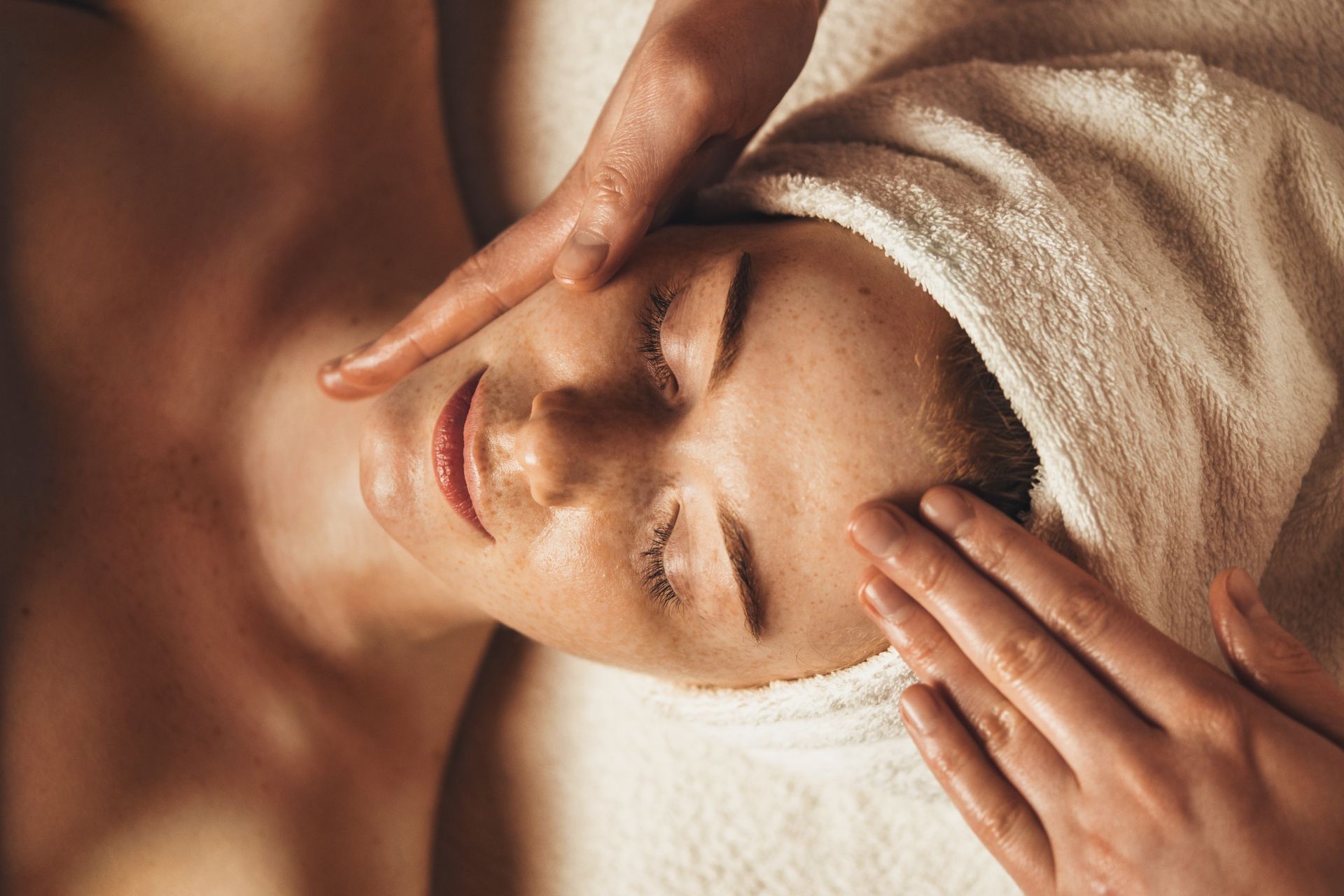 Woman receiving a facial massage with hands on forehead. She is relaxed with eyes closed, and wearing a towel.