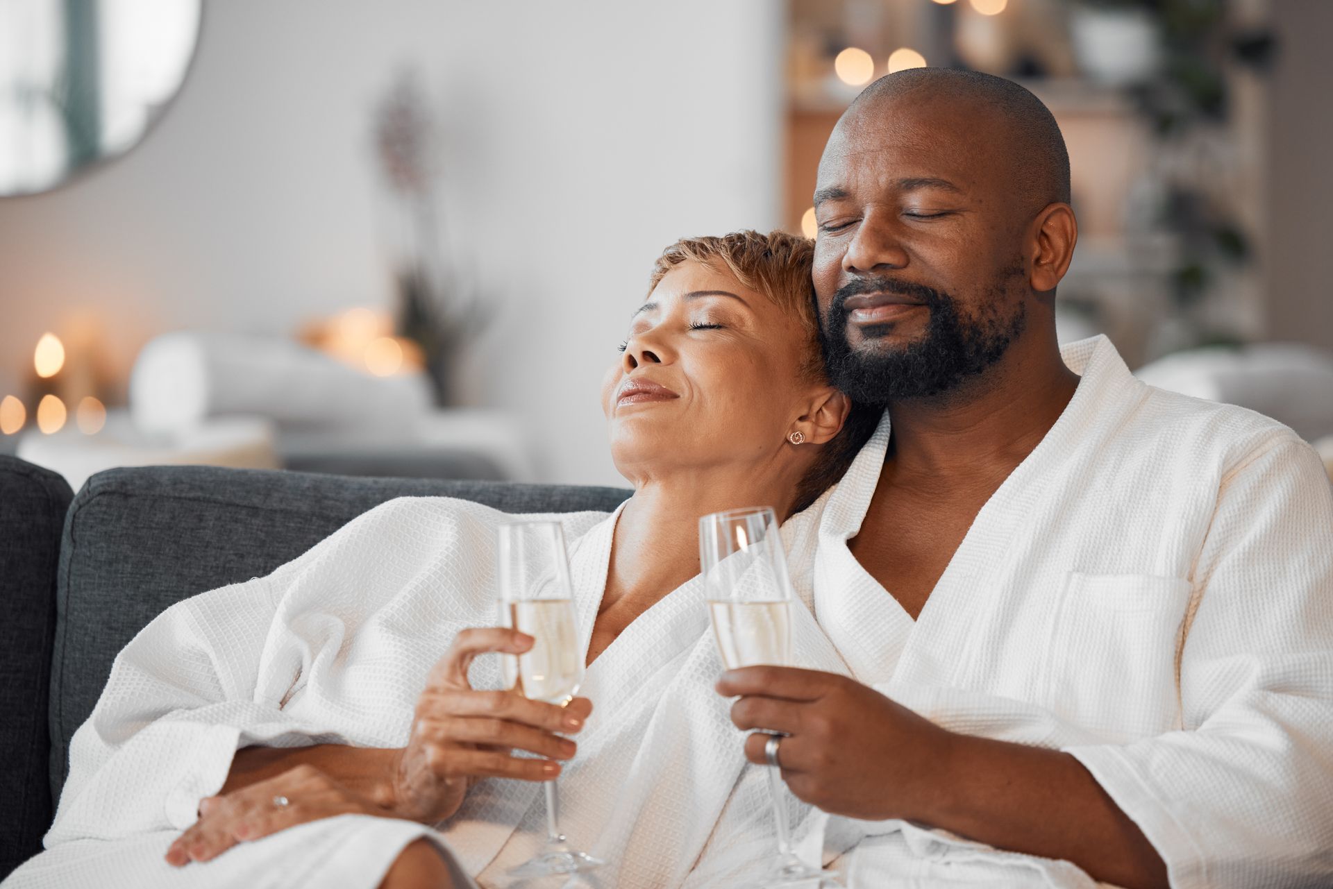 Couple in bathrobes relax with champagne, eyes closed, in a spa-like setting.