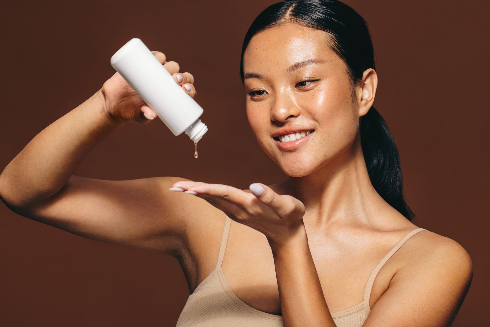 Woman smiling, pouring skincare product into hand against brown backdrop.