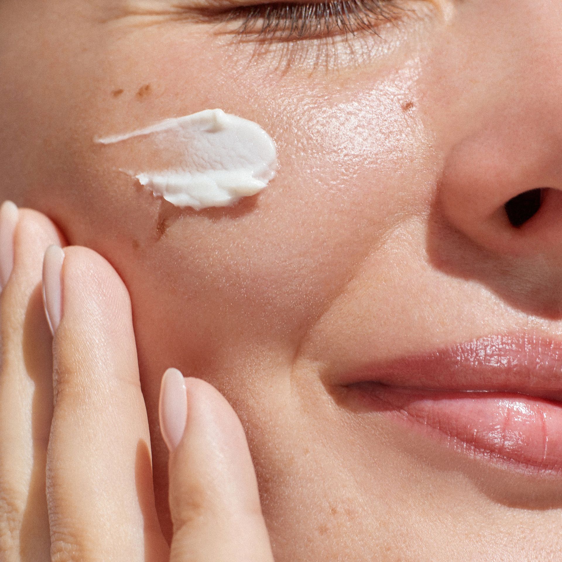 A close-up of a person applying a dab of white skincare cream to their cheek with their hand.