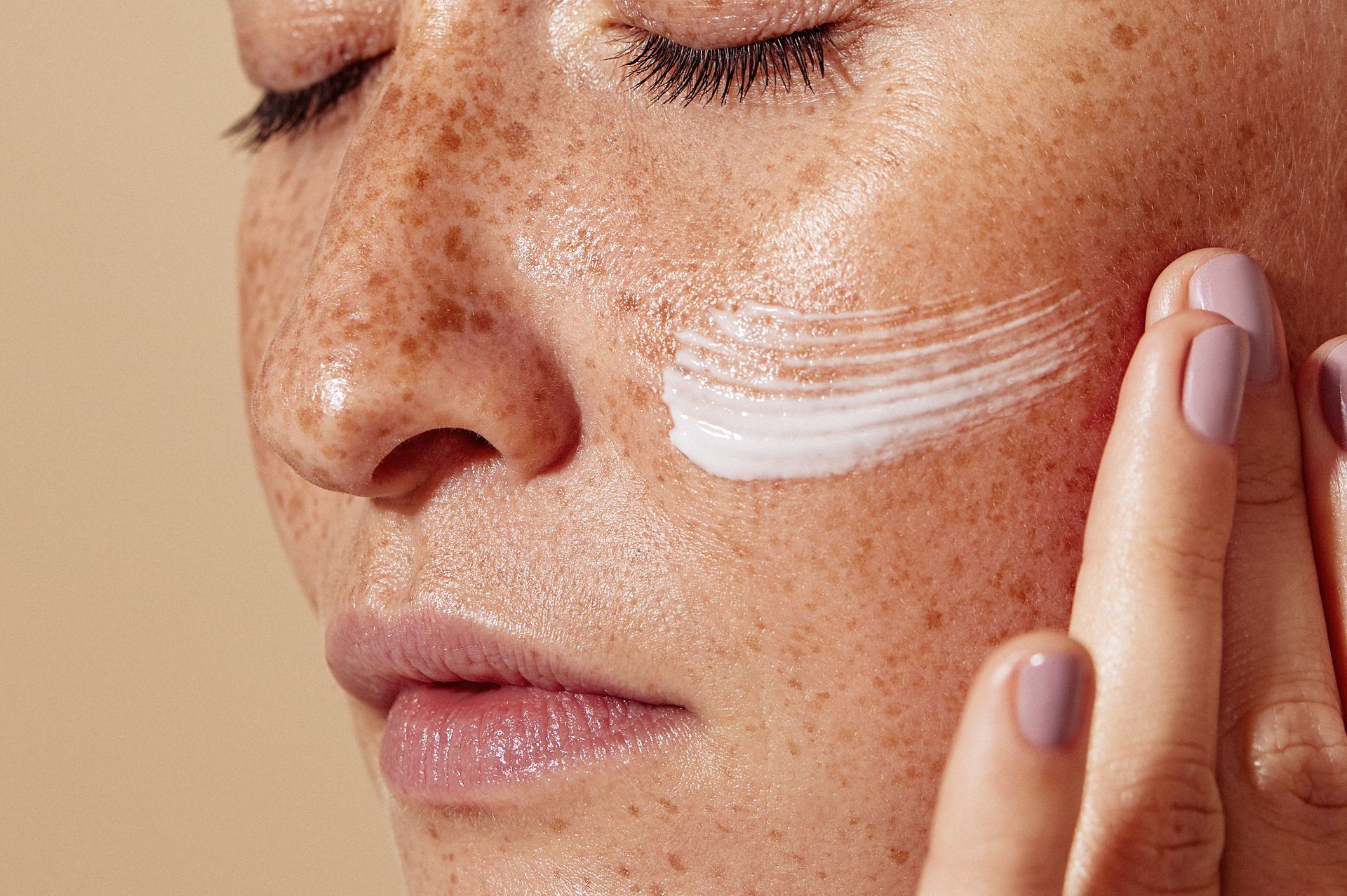 Close-up of a person applying white face cream to their cheek with eyes closed