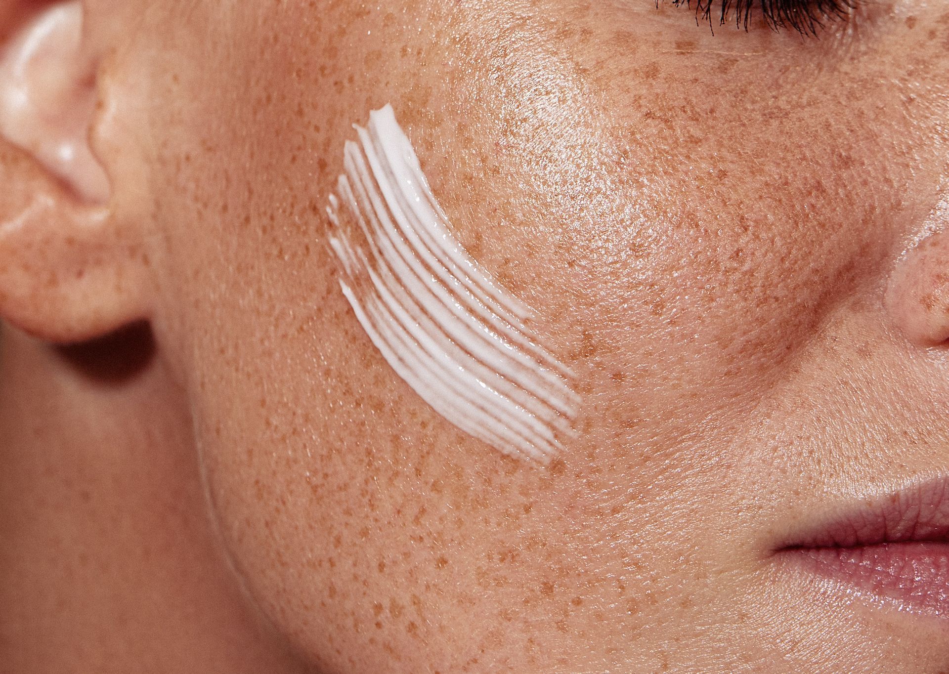 Close-up of a face with a white cream swatch on the cheek, showing skin texture and makeup application