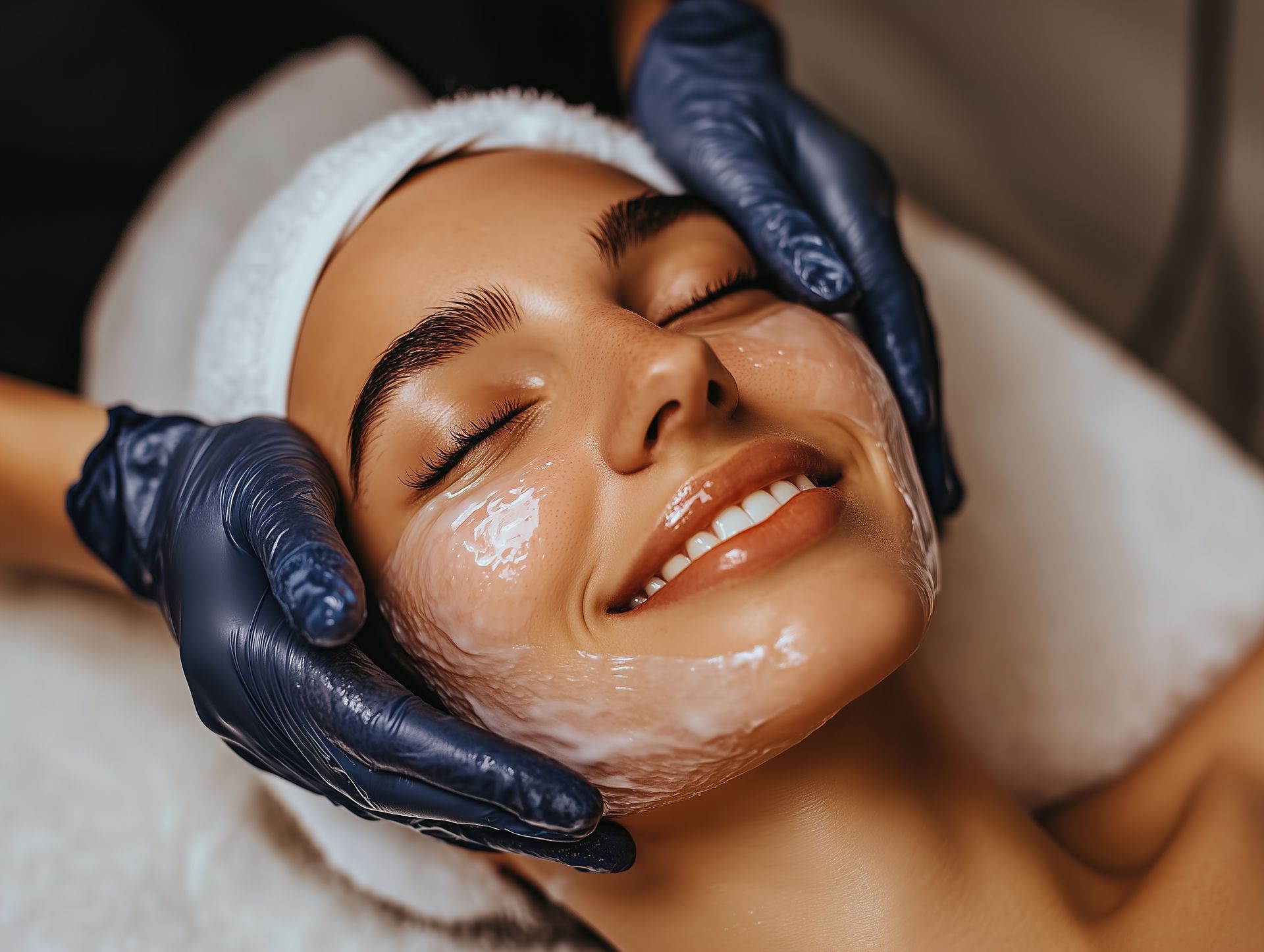 Woman receiving facial treatment, smiling with eyes closed, hands applying cream, wearing a white headband.