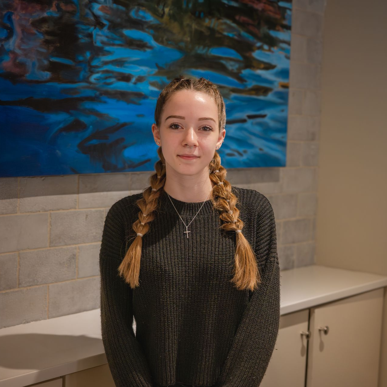 Woman with braided hair stands in front of a blue art piece, wearing a gray sweater.