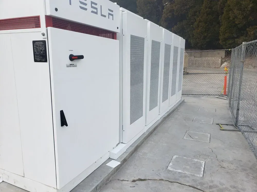 A large white tesla battery is sitting next to a fence.