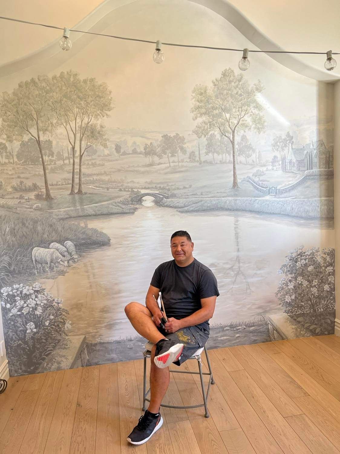 A man is sitting on a stool in front of a painting of a river.