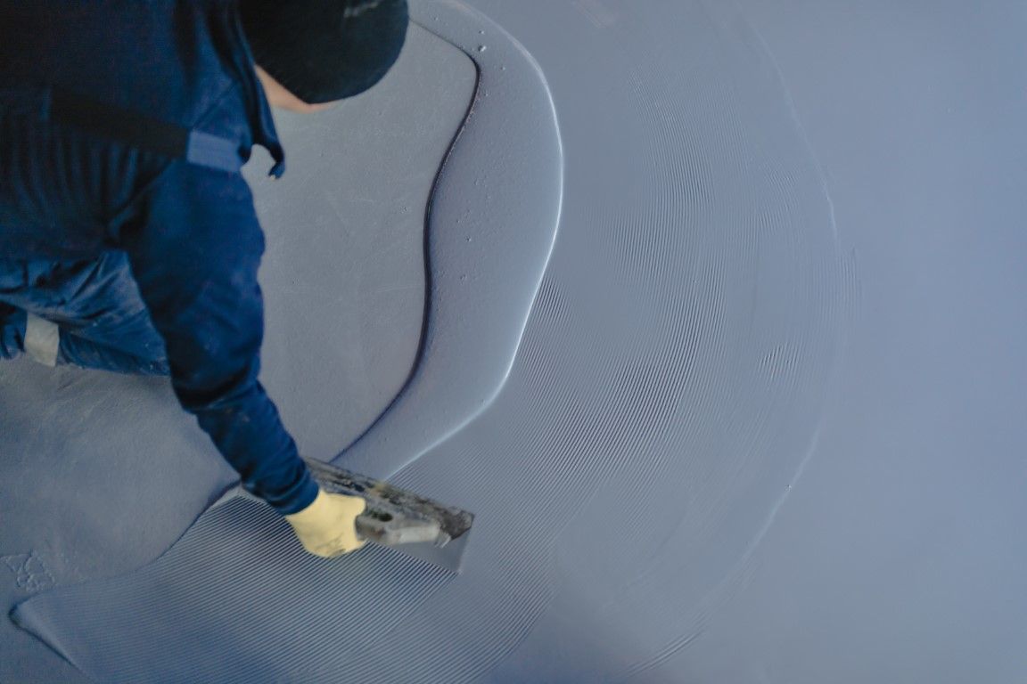 Person in blue coveralls applying adhesive to a floor with a notched trowel.