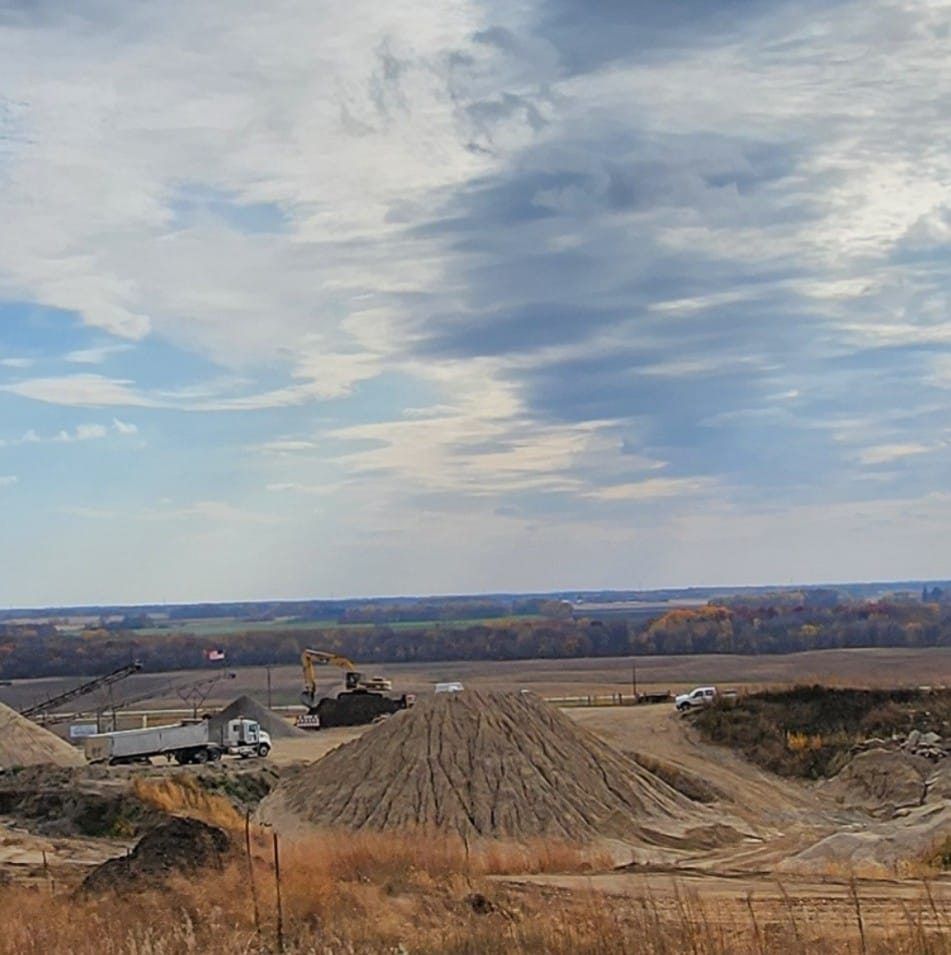 Construction site with excavator and trucks on a dirt road under a cloudy sky, overlooking fields