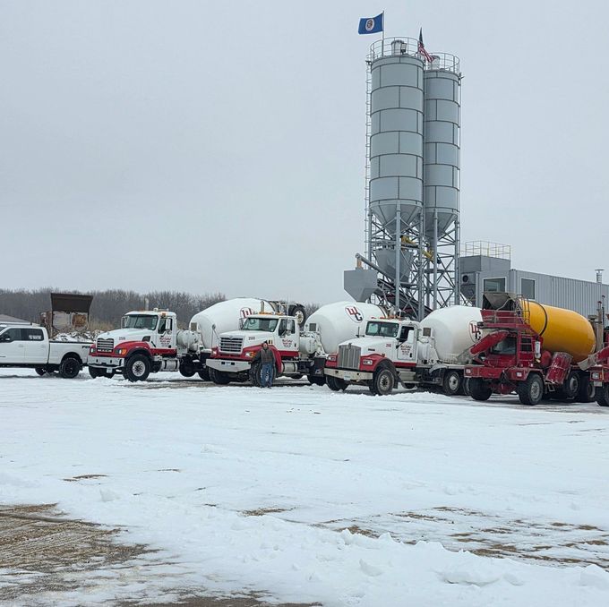 Concrete mixer trucks lined up in a snowy industrial yard beside a tall silo under an overcast sky