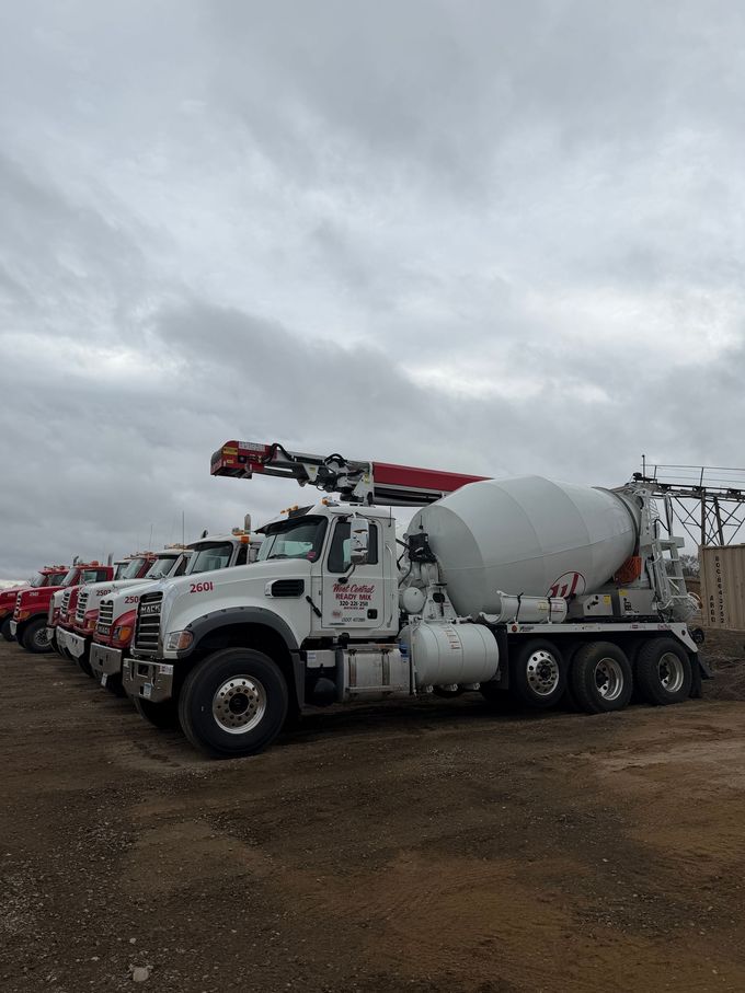 White concrete mixer truck with red crane parked on a muddy lot under cloudy skies