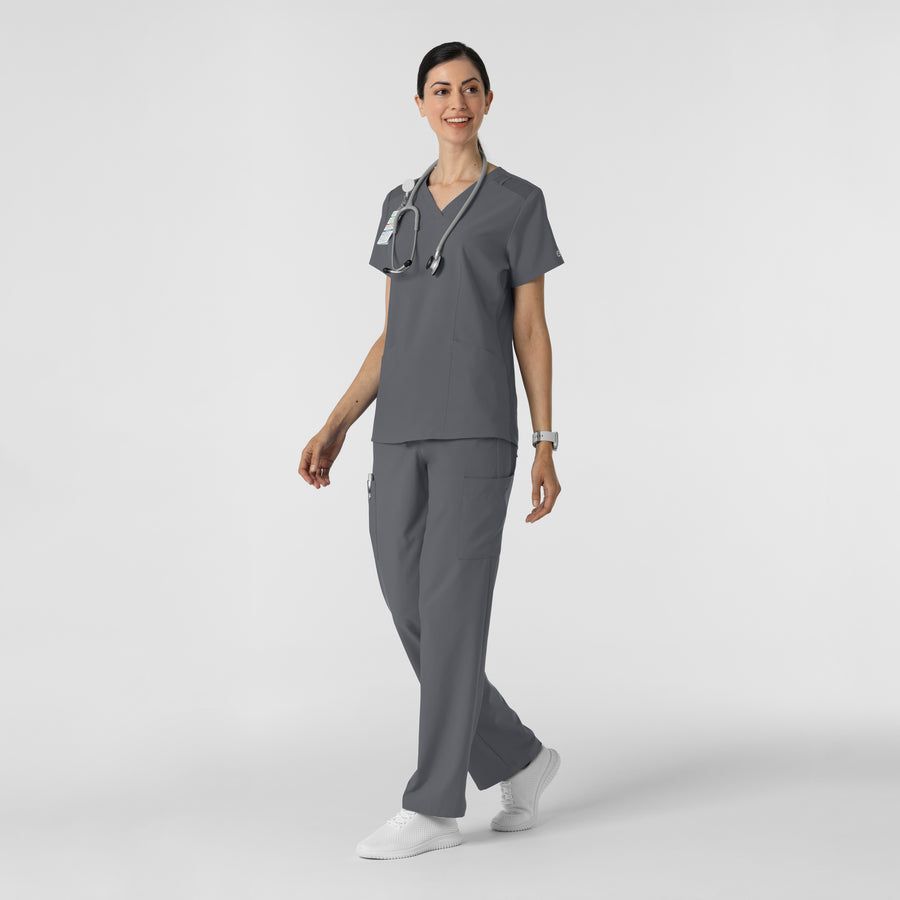 Woman in grey scrubs walks, smiles, stethoscope around neck.