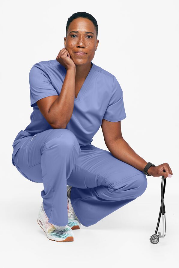 Person in blue scrubs squats, holding a stethoscope. She rests her chin on her hand and smiles.
