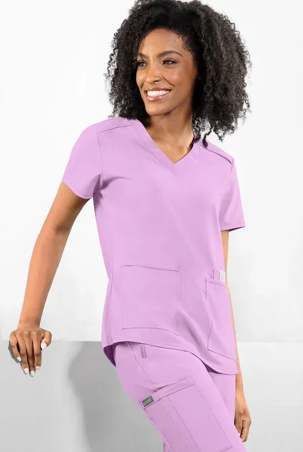 Woman in lavender scrubs, leaning against a white surface, smiling.