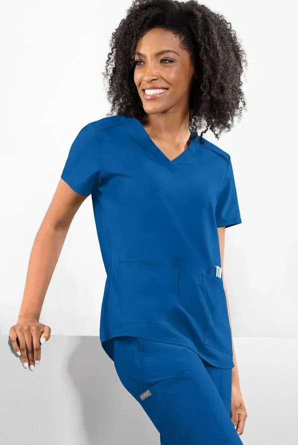 Woman wearing blue scrubs smiles, leans against a white surface, indoors.