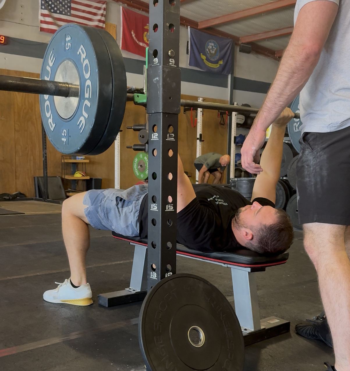 Muscular man sits shirtless, hands clasped, serious expression. Dark setting, black and white photo.