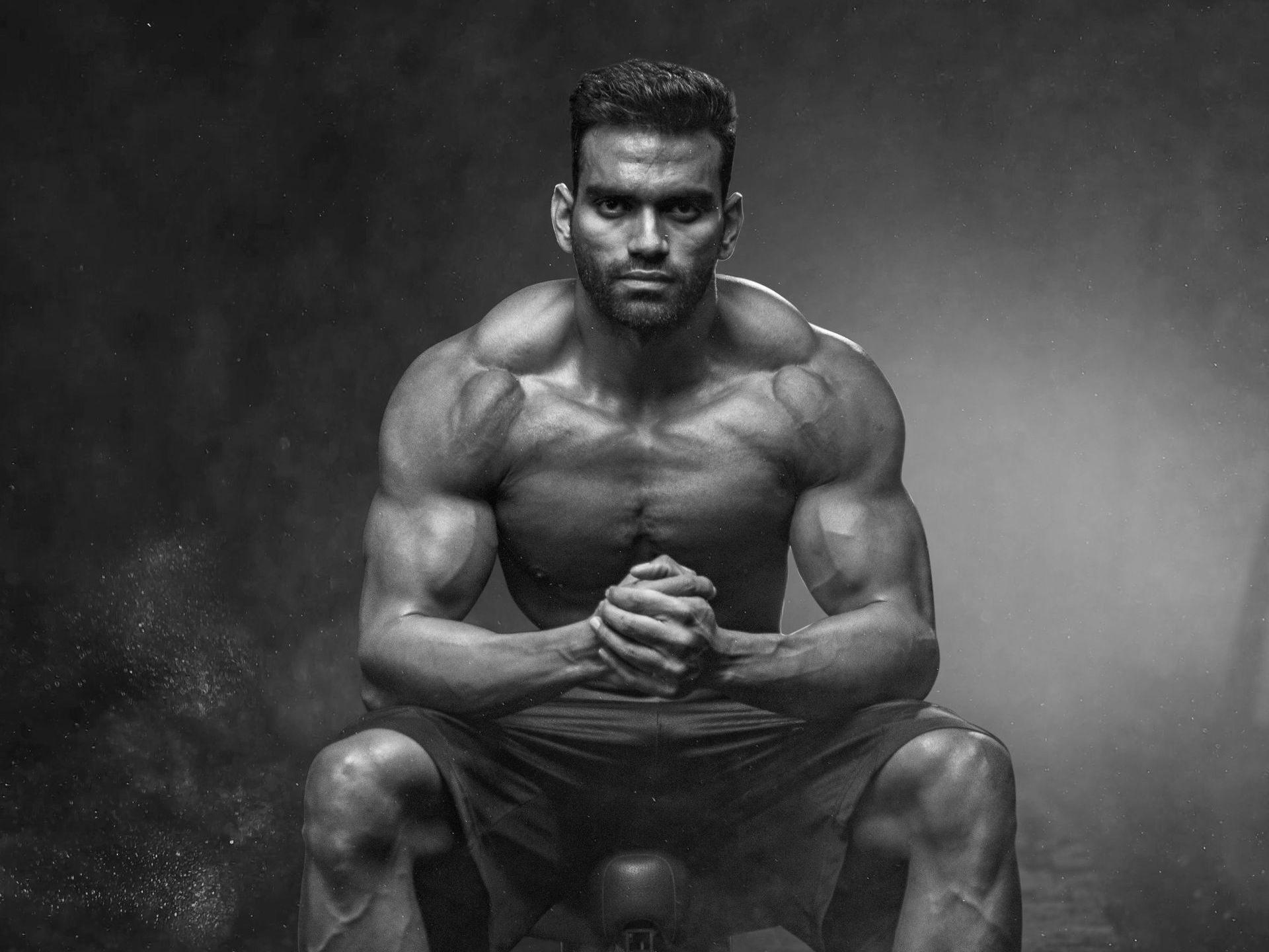 Muscular man sits shirtless, hands clasped, serious expression. Dark setting, black and white photo.