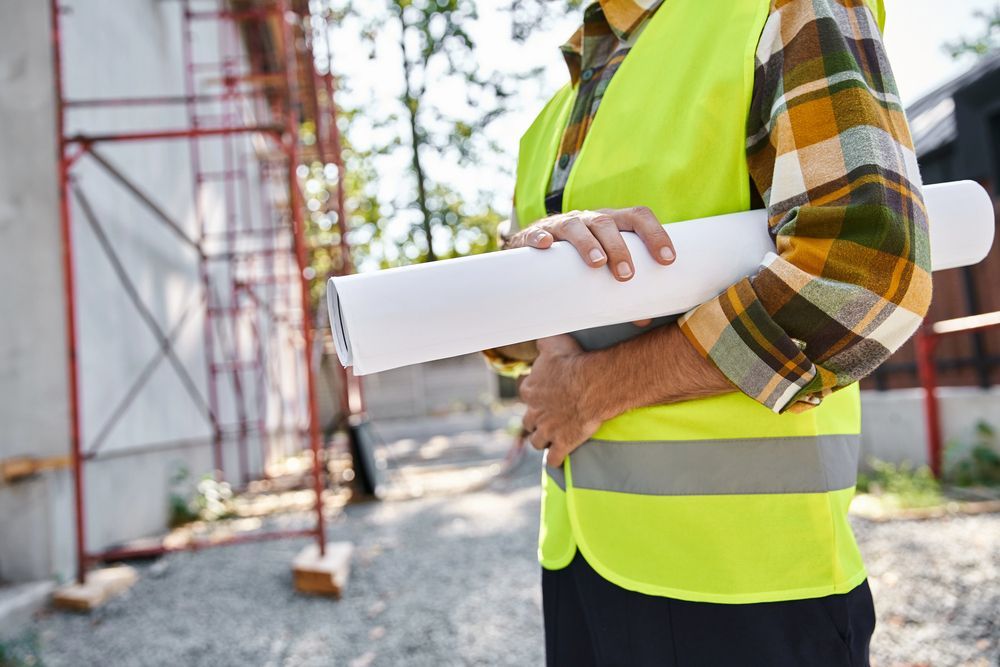 Un operaio edile con un giubbotto di sicurezza giallo tiene in mano delle planimetrie arrotolate all'esterno di un edificio in costruzione.