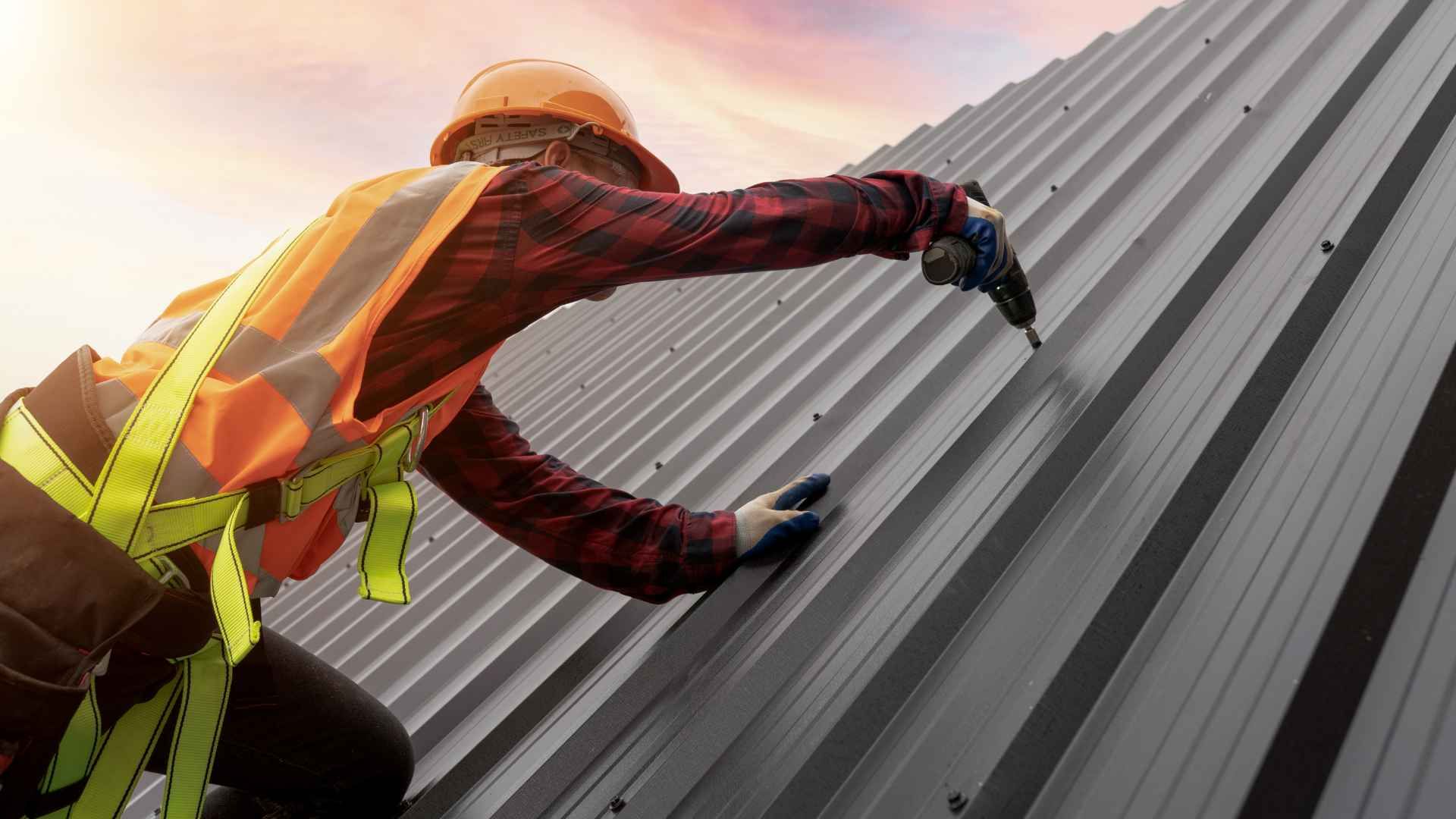A person in safety gear is using a power drill to install or secure a metal roof panel.
