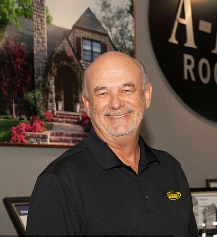 A man in a black shirt is smiling in front of a picture of a house