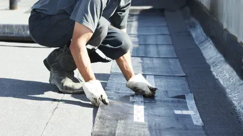 A man is kneeling down and applying a roof coating to a roof.