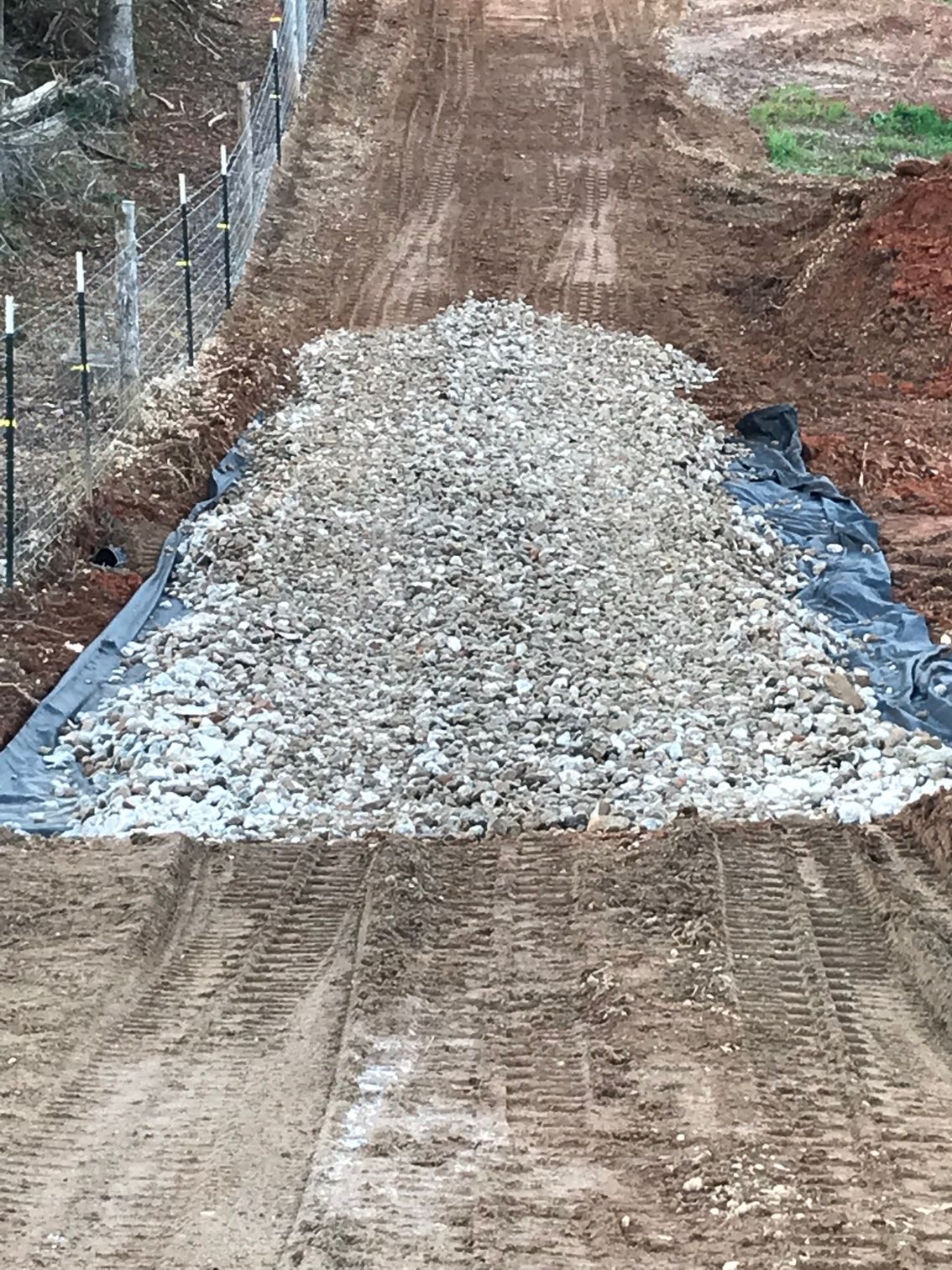 Pile of gravel on black fabric covering dirt road; fence on left.