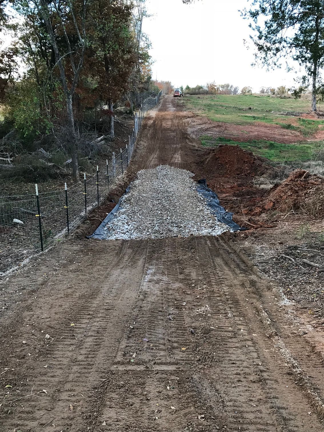 Dirt road with gravel section, bordered by a wire fence and dirt mounds, leading uphill through a rural area.