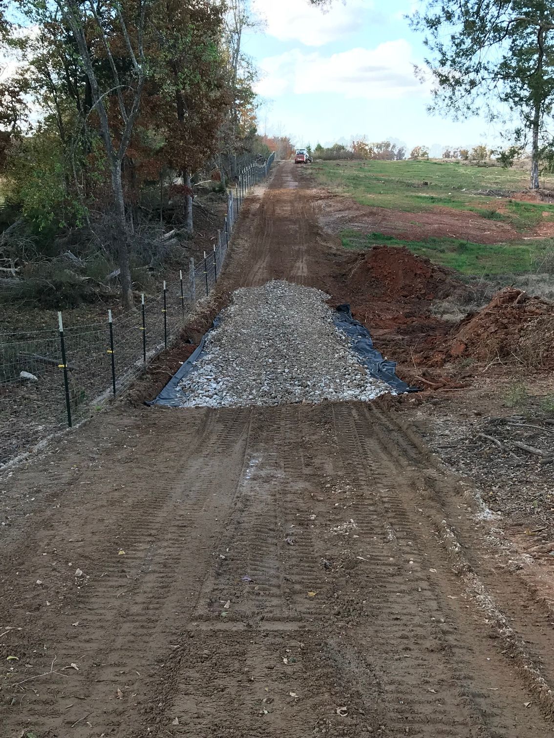 Dirt road leading uphill, transitioning to gravel. Fence borders the left side, and trees are visible.