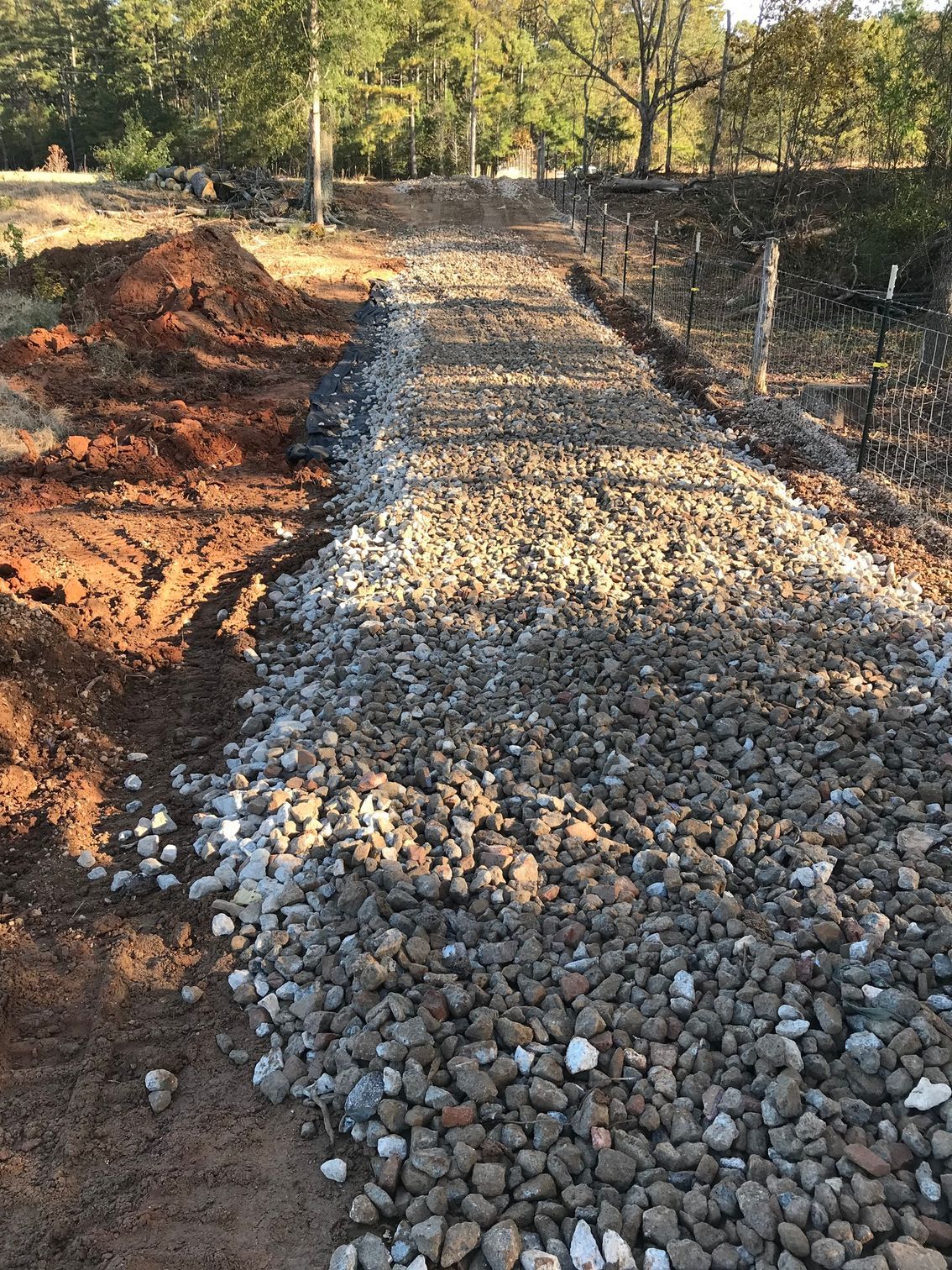 Gravel path under construction with red dirt pile on the left, a fence on the right, and trees in the background.