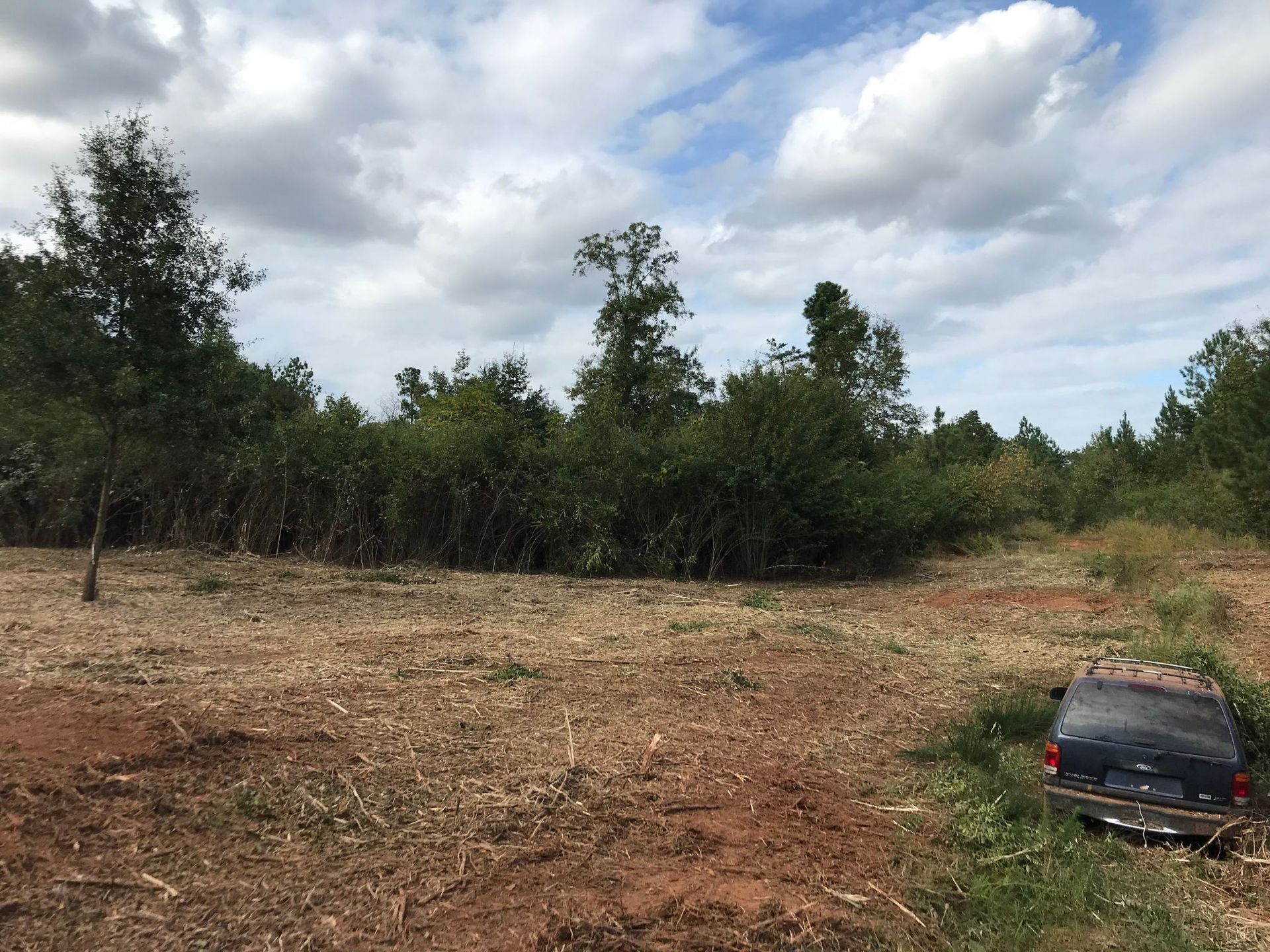 Cleared land with a rusted vehicle in the right corner and a treeline in the background under a cloudy sky.