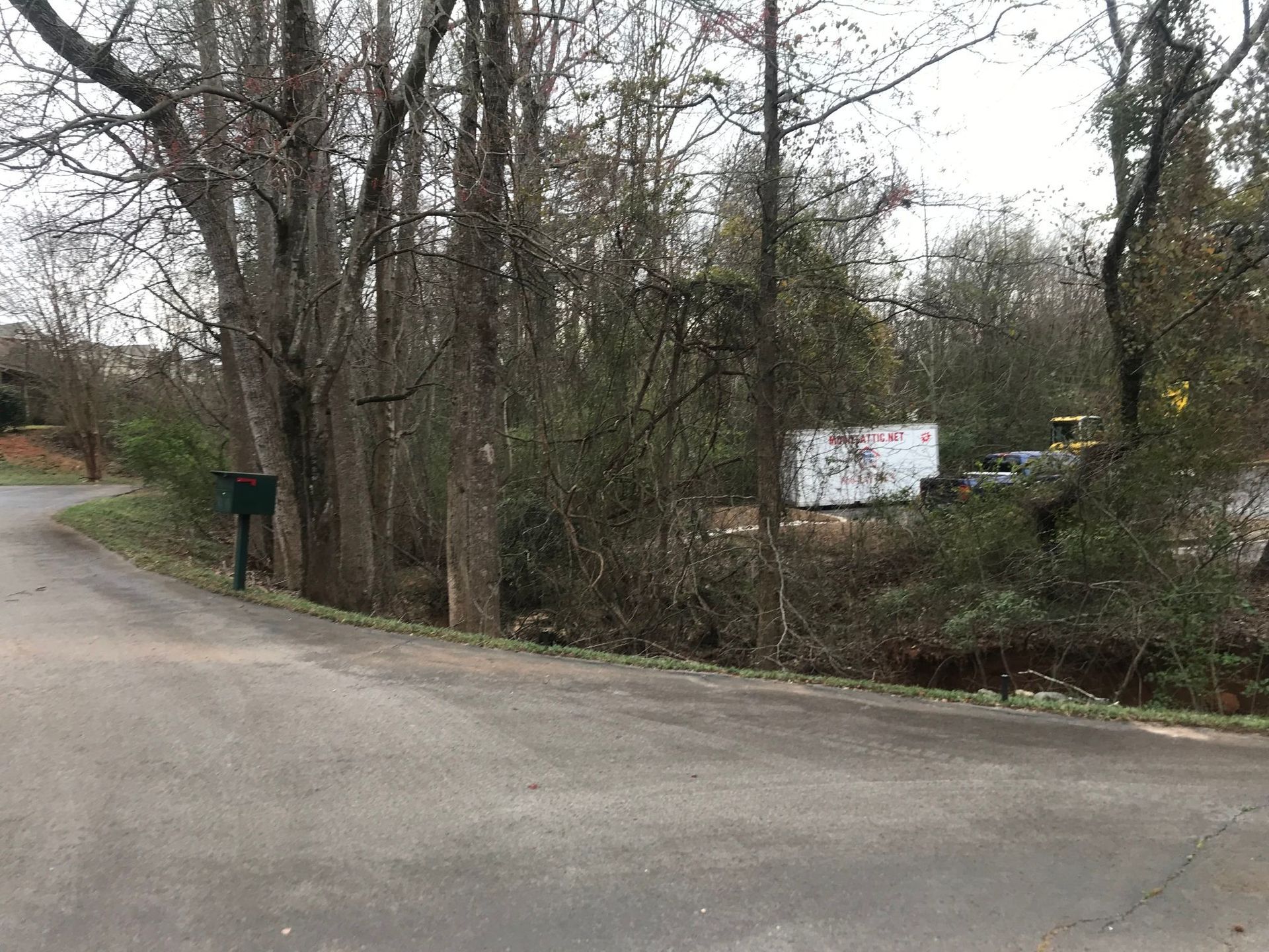 A paved road curves past a tree-lined area with a mailbox, leading to a white building in the distance.
