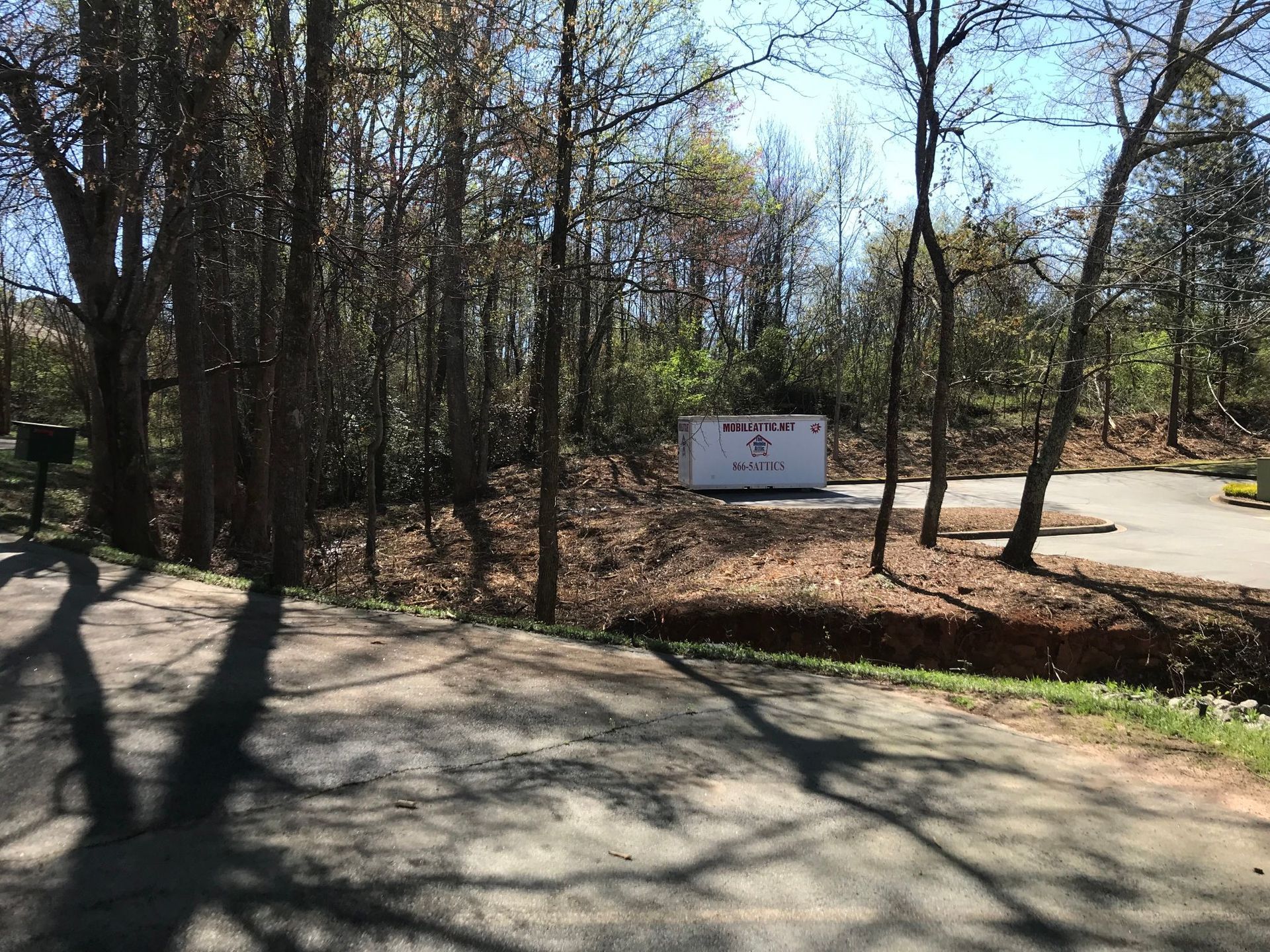 A white utility box sits in a wooded area beside a road, under a sunny sky.