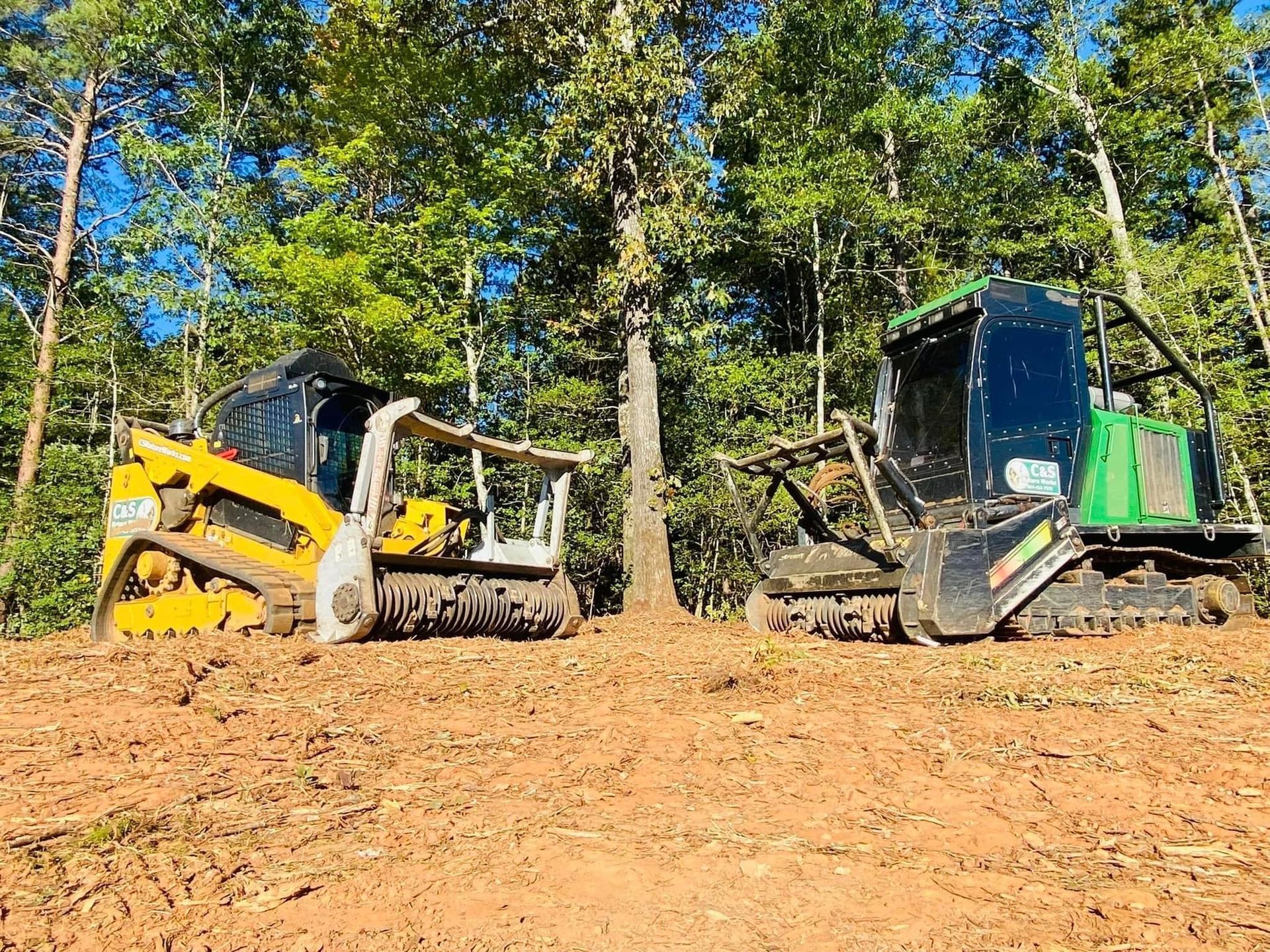 Two forestry mulchers on a cleared dirt area, tree in between them, surrounded by trees.