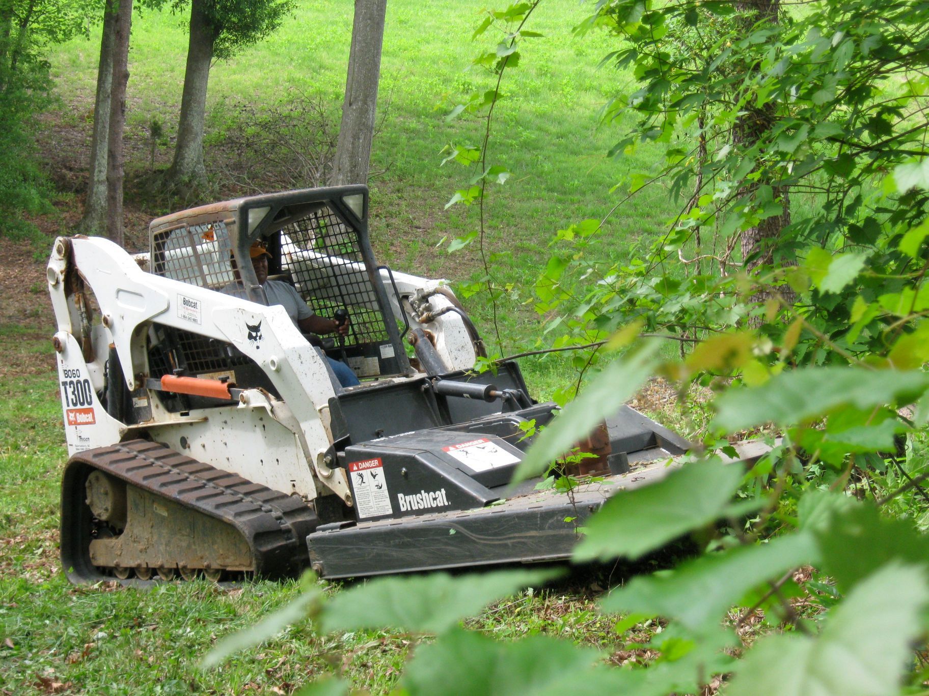Bobcat skid-steer with mower attachment cutting brush near trees on grassy hillside.